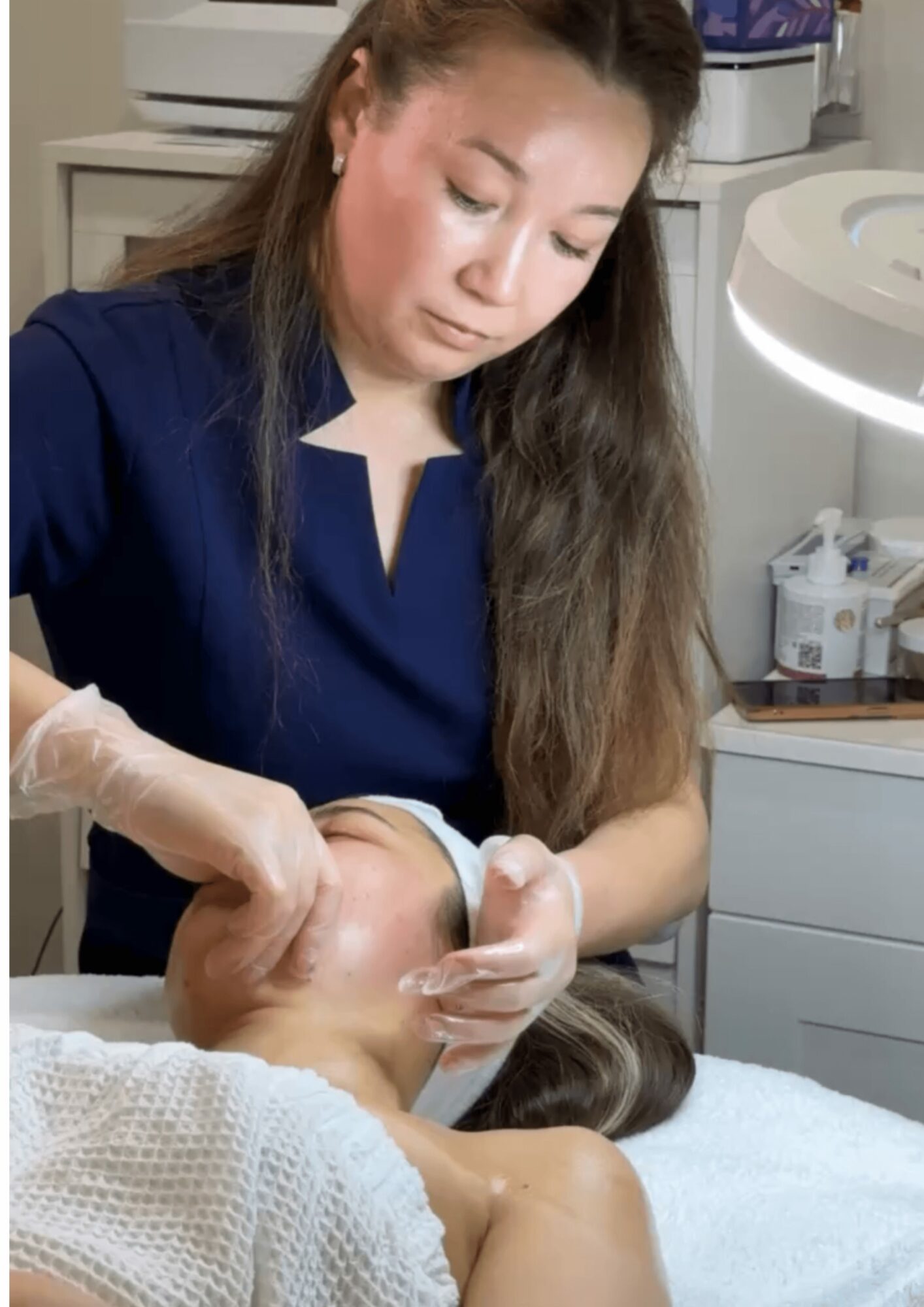 A woman in a dark blue uniform performs a facial treatment on a person lying down, in a clinical setting.