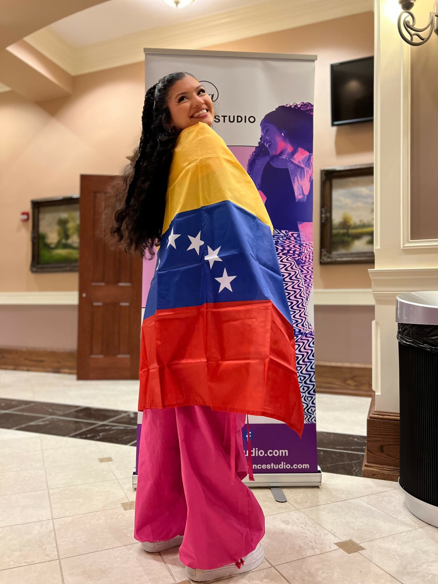 Woman standing indoors, wrapped in a Venezuelan flag, smiling, with paintings and a TV in the background.