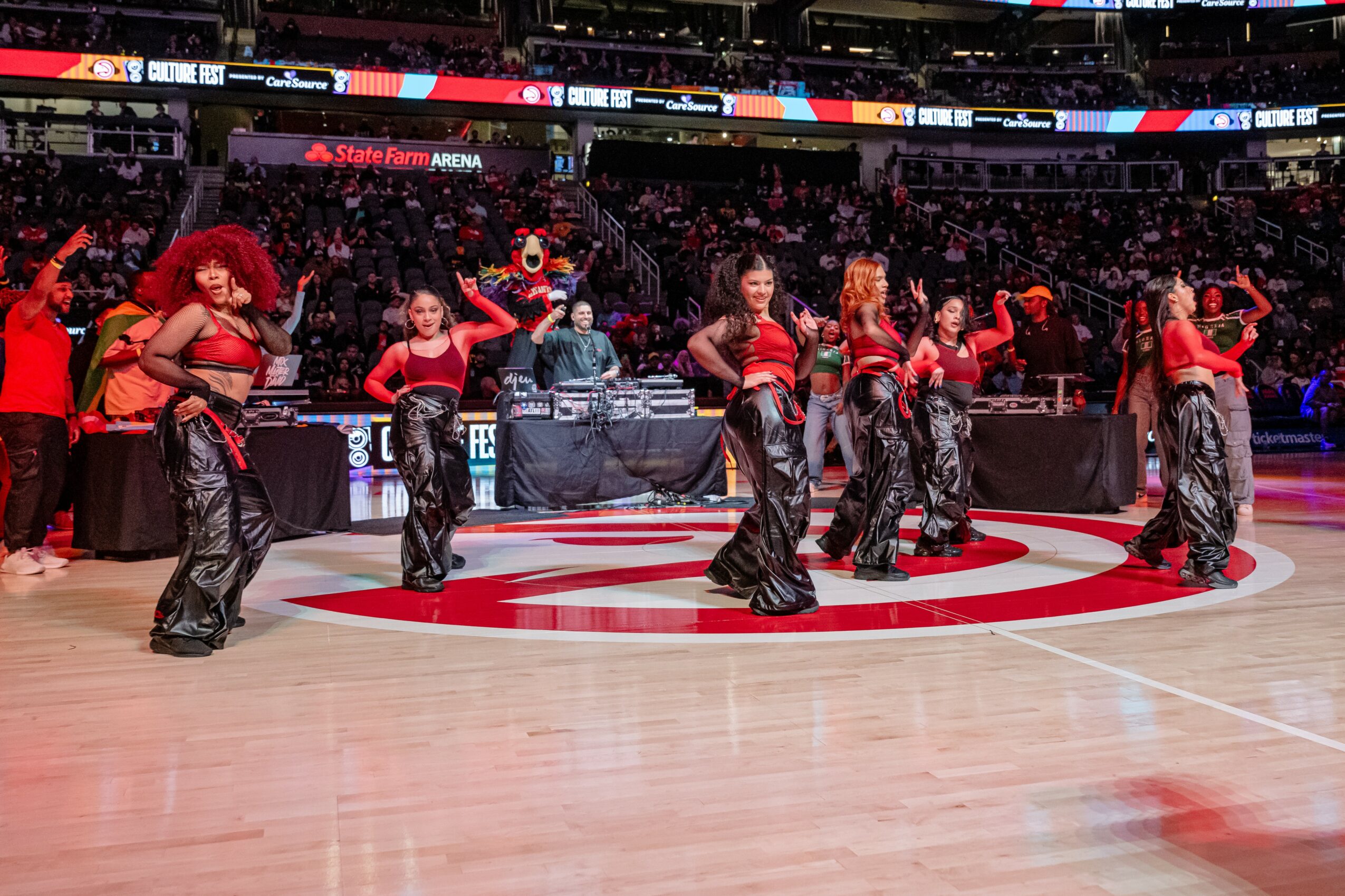 Group of performers dancing on basketball court with audience in background, some wearing red tops and black pants.