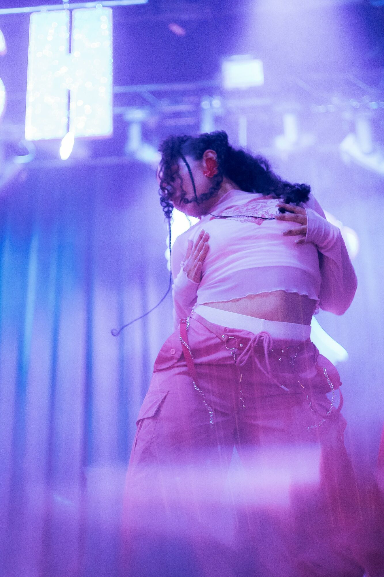 Person with curly hair dancing in a pink outfit under purple and blue stage lights.