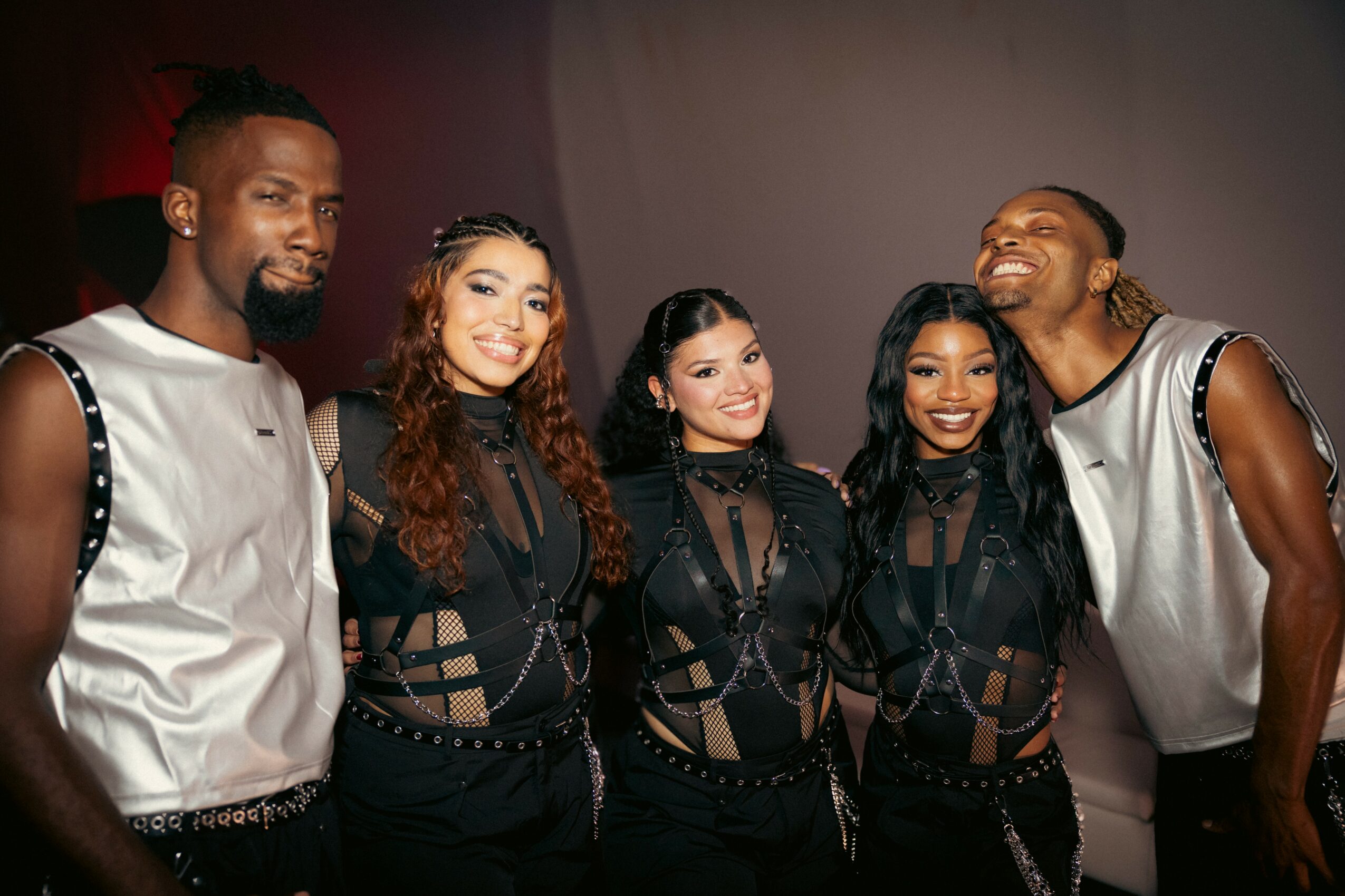 Group of five people smiling, standing close together, wearing black outfits with sheer details, against a plain background.