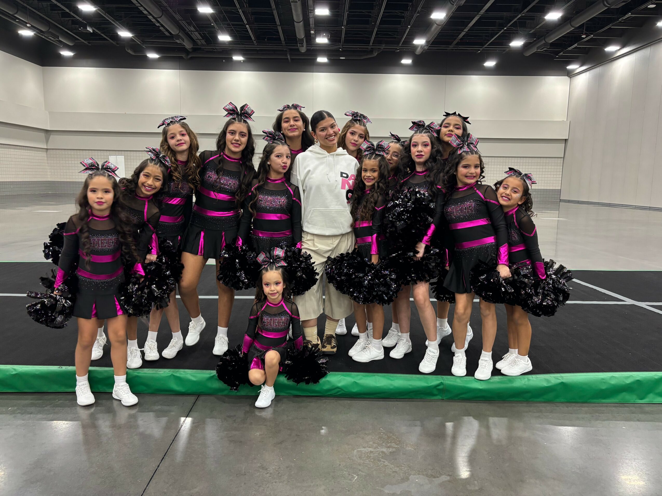 Group of young cheerleaders in black and pink uniforms with pom-poms, posing indoors on a green mat.