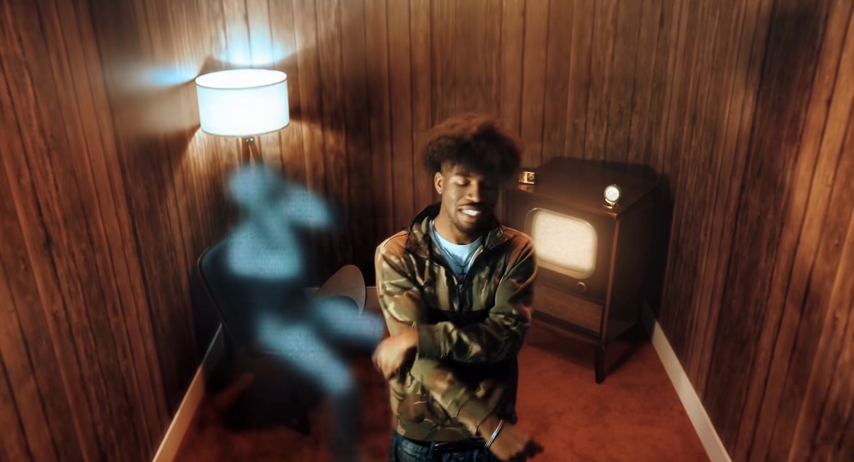 Young man with curly hair standing in a small room with wooden walls, illuminated by a lamp, with a blurred figure nearby.