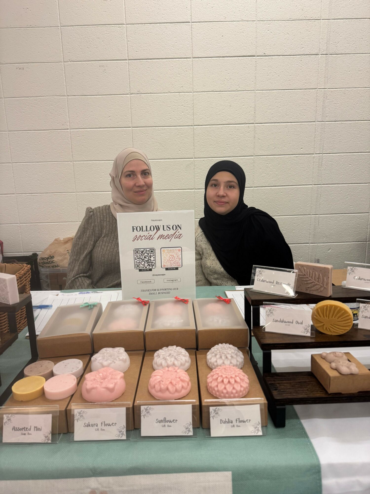 Two women sit behind a table with baked goods and a sign, against a white tiled wall.