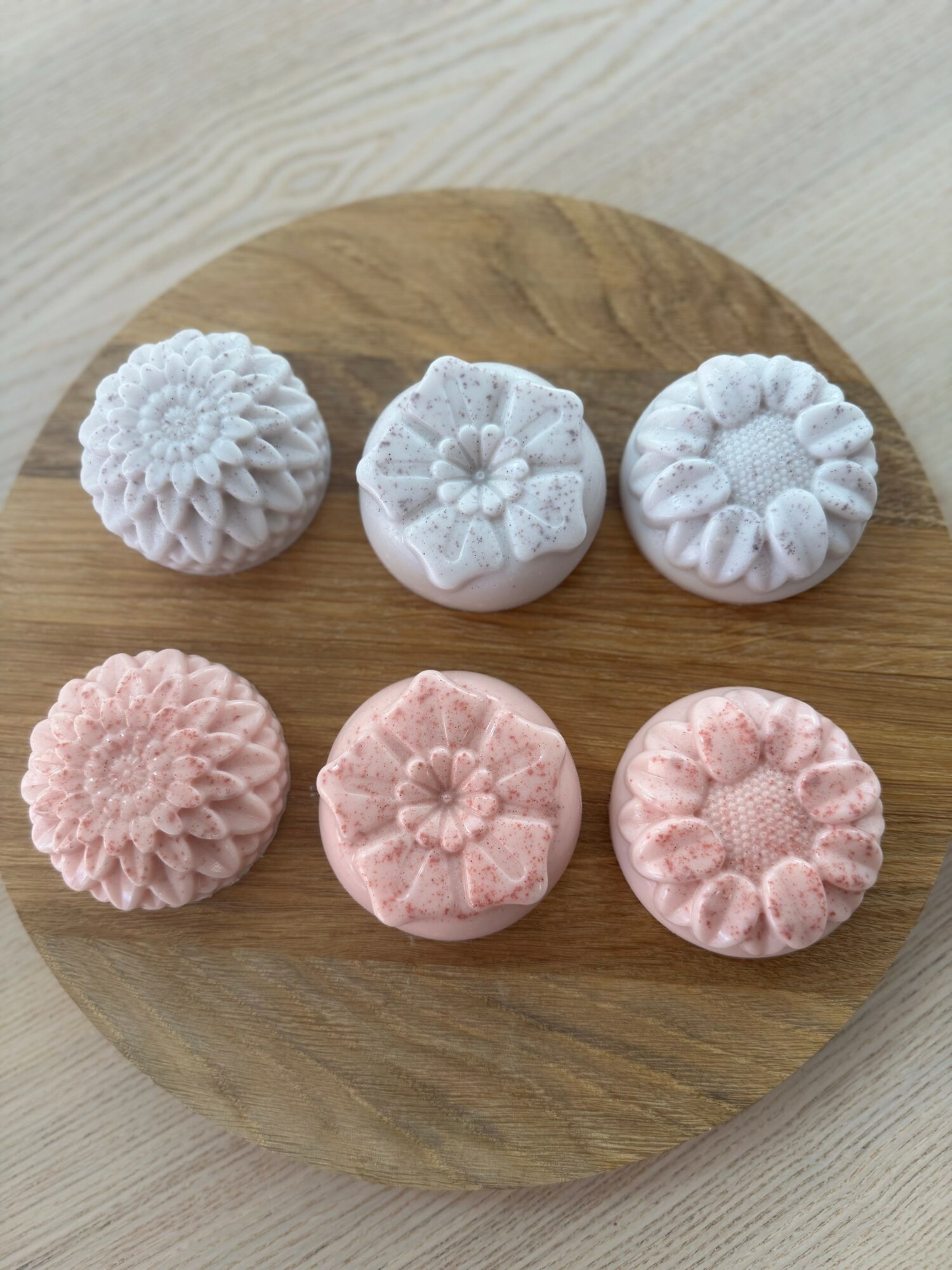 Six intricately carved soap bars in white and pink, arranged on a round wooden board.
