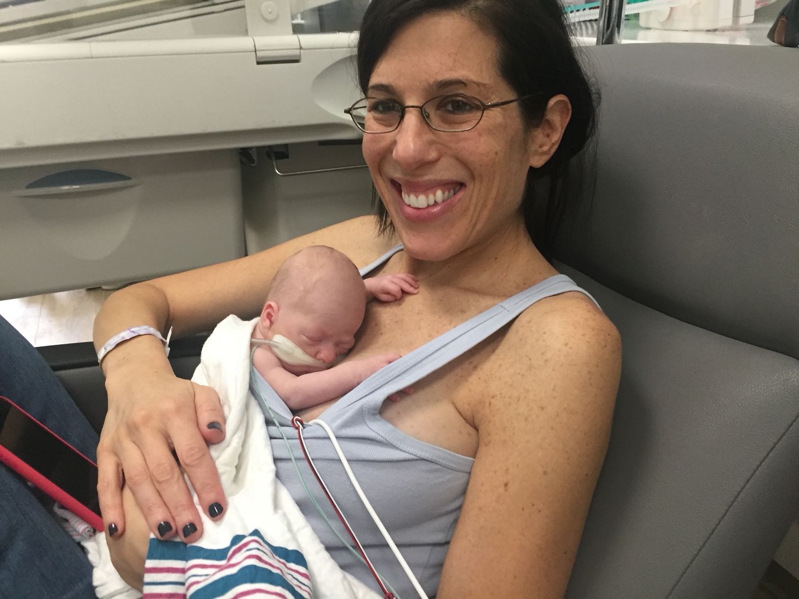 Woman with glasses holding a newborn baby in a hospital room, smiling at the camera.