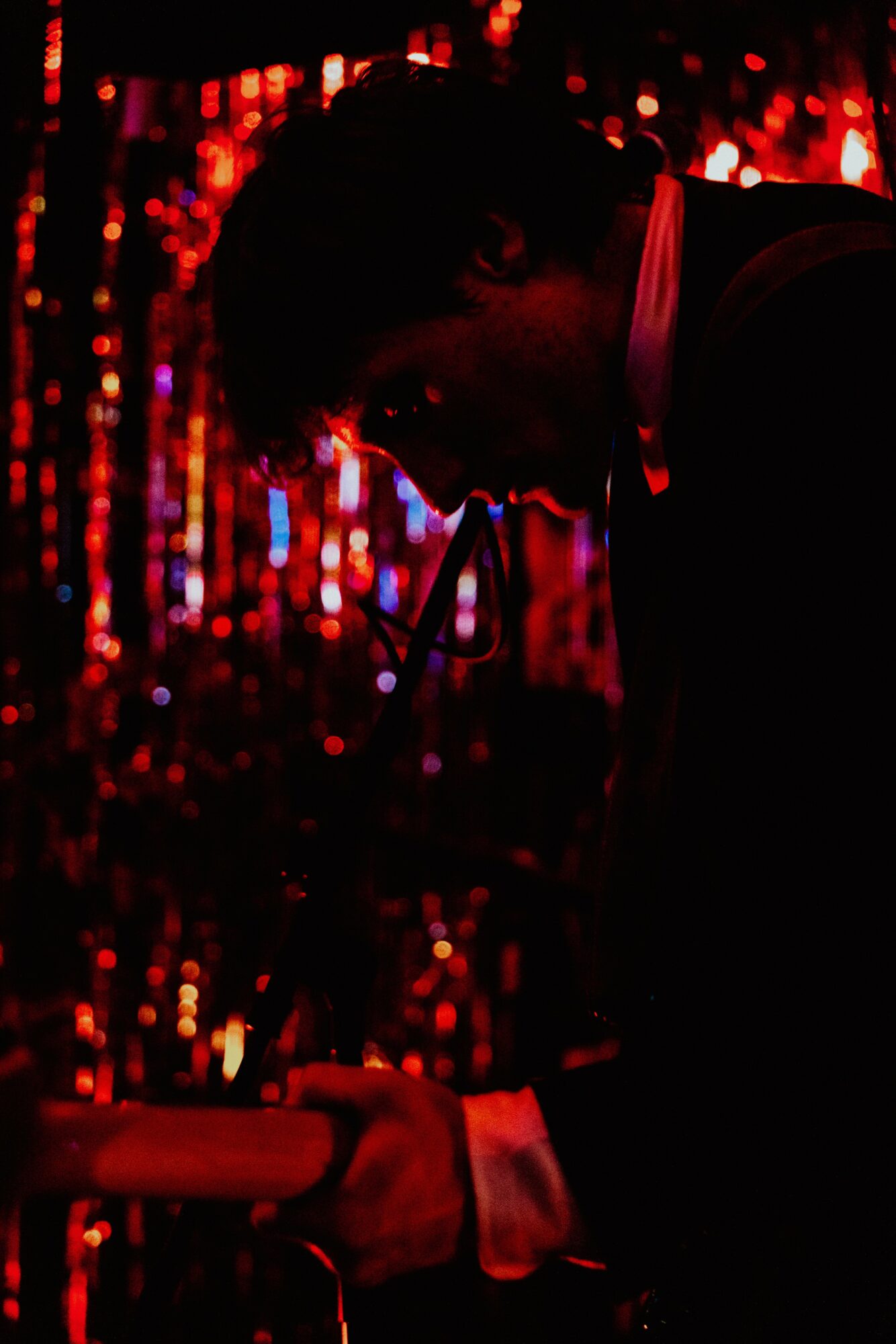 Person with dark hair in a dark setting, holding an object, illuminated by red and purple lights in the background.
