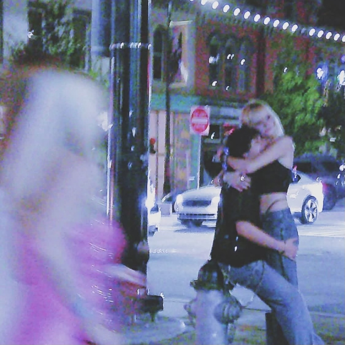 Two people hugging on a city street at night, with cars and buildings in the background.
