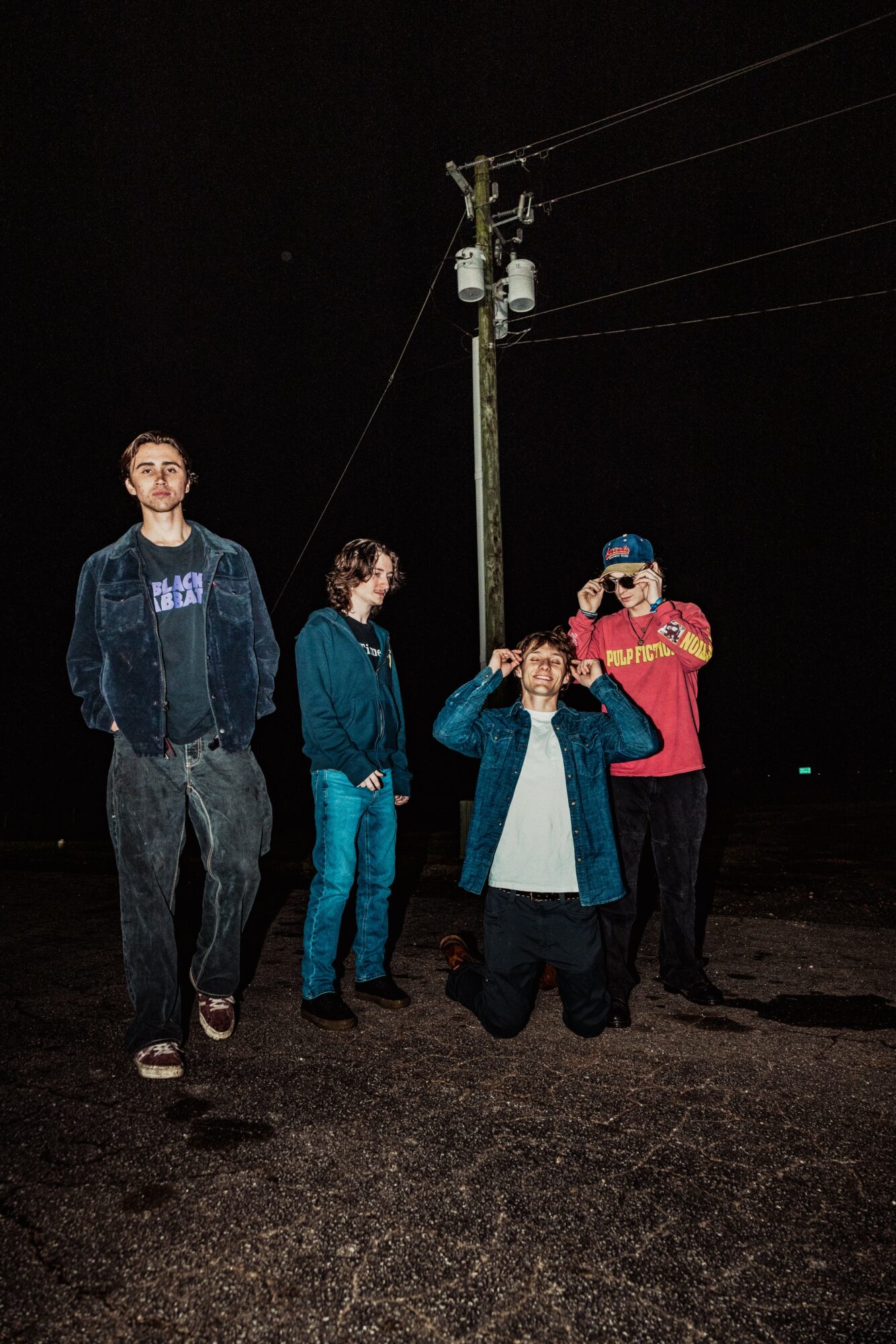 Five young people standing outdoors at night near a utility pole, some wearing jackets and sunglasses, one kneeling.