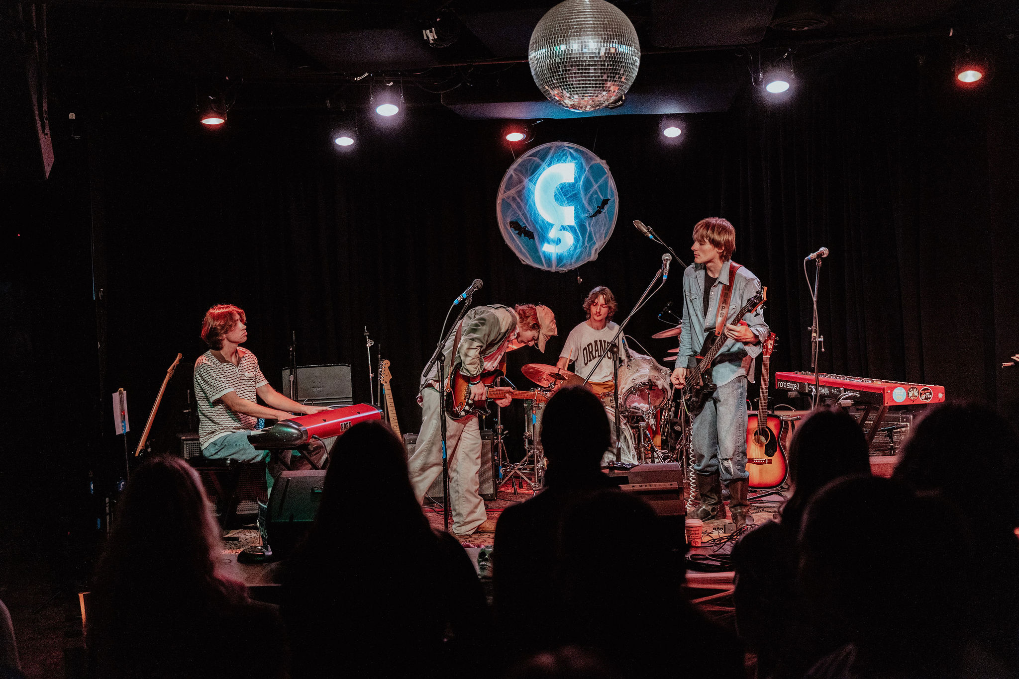 Band performs on stage with four members, musical instruments, and a disco ball overhead, audience silhouettes in foreground.
