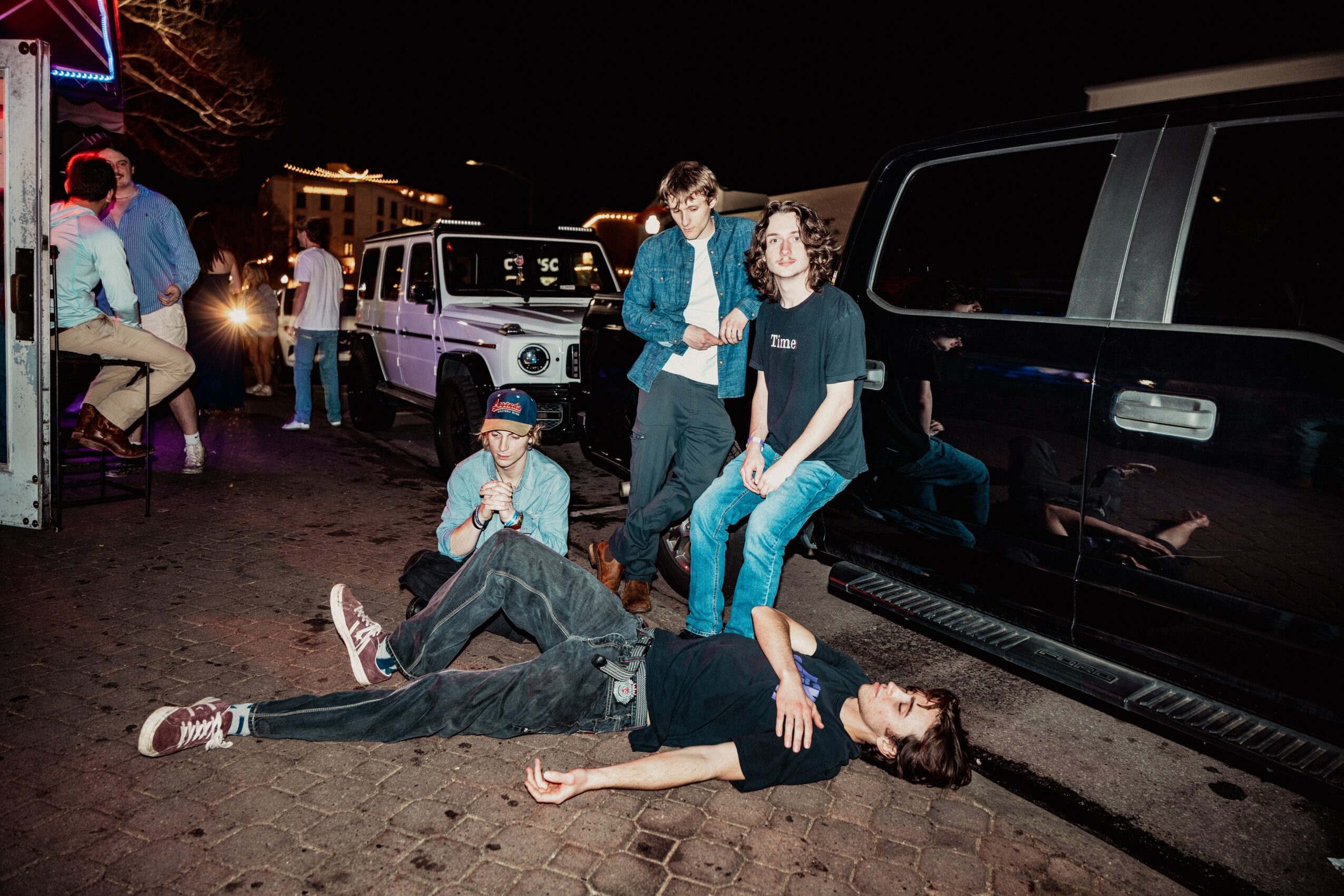 Person lying on the ground with others standing nearby at night, vehicles in the background.