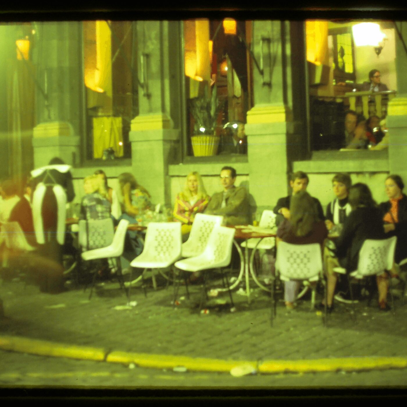 People sitting at tables outside a cafe or restaurant, with large windows and yellow lighting inside.