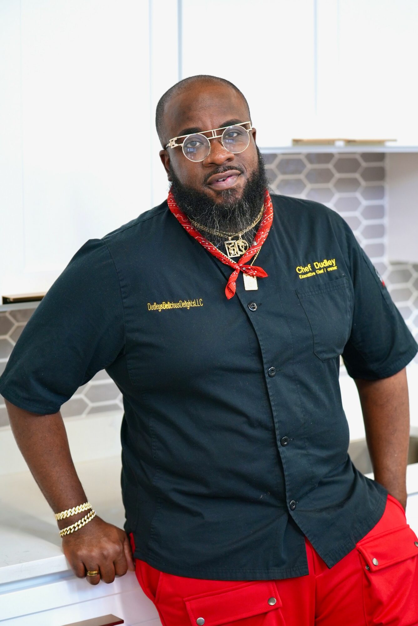 Man with glasses, beard, and jewelry wearing a black shirt and red pants indoors.