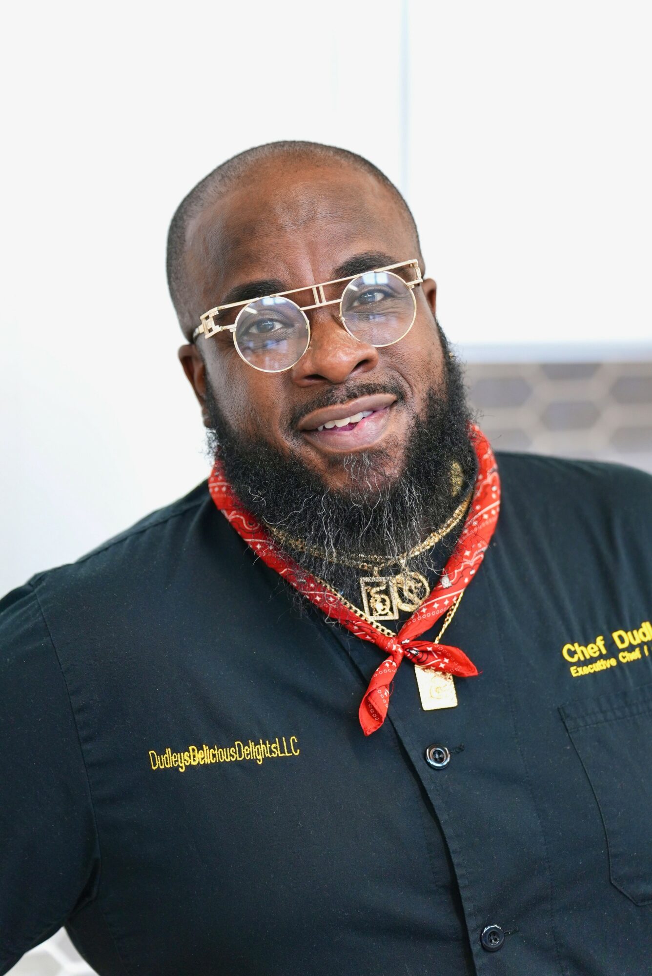 Man with glasses, beard, wearing a black shirt with gold embroidery, red bandana, and gold necklace.