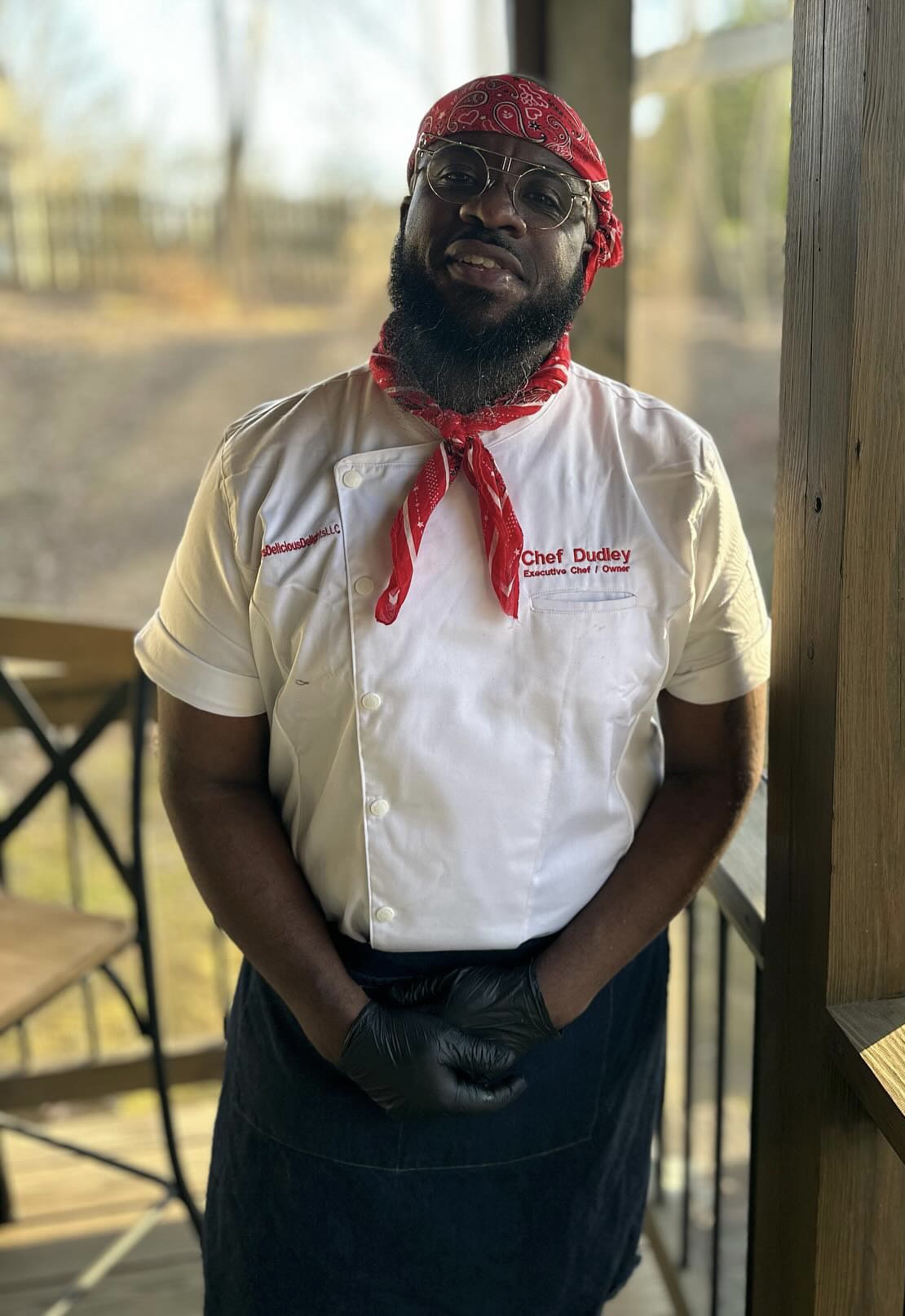 Person wearing chef's uniform, red bandana, sunglasses, standing outdoors with trees in background.