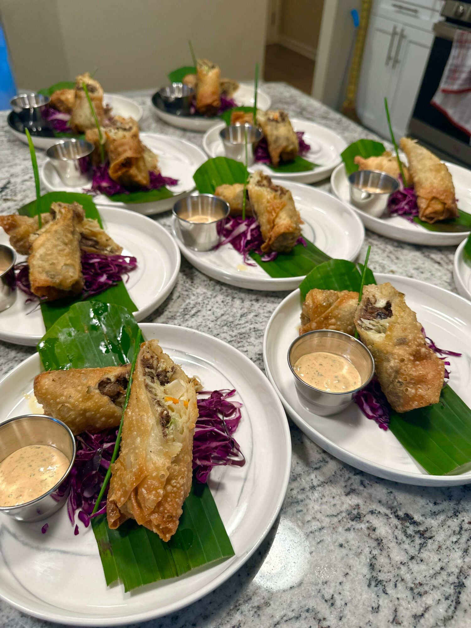 Multiple plates with fried spring rolls, purple cabbage, green leaf, and dipping sauce on a marble table.