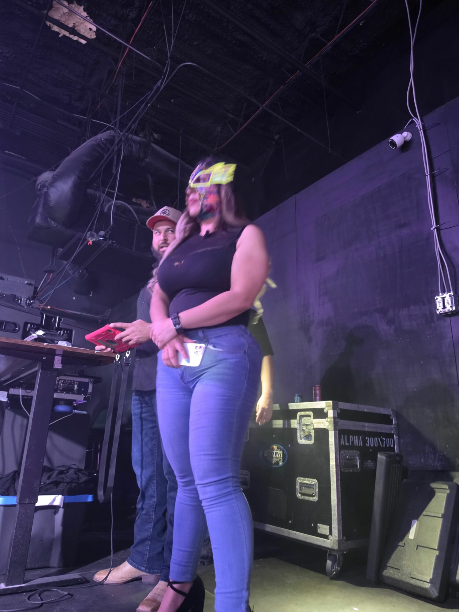 Person wearing a helmet and headphones standing next to another person in a dark room with equipment and cables.