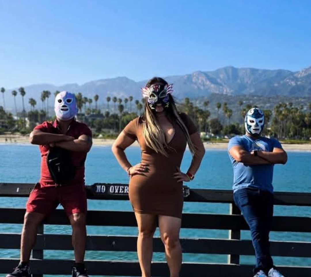 Three people wearing animal masks stand on a pier with water and mountains in background.
