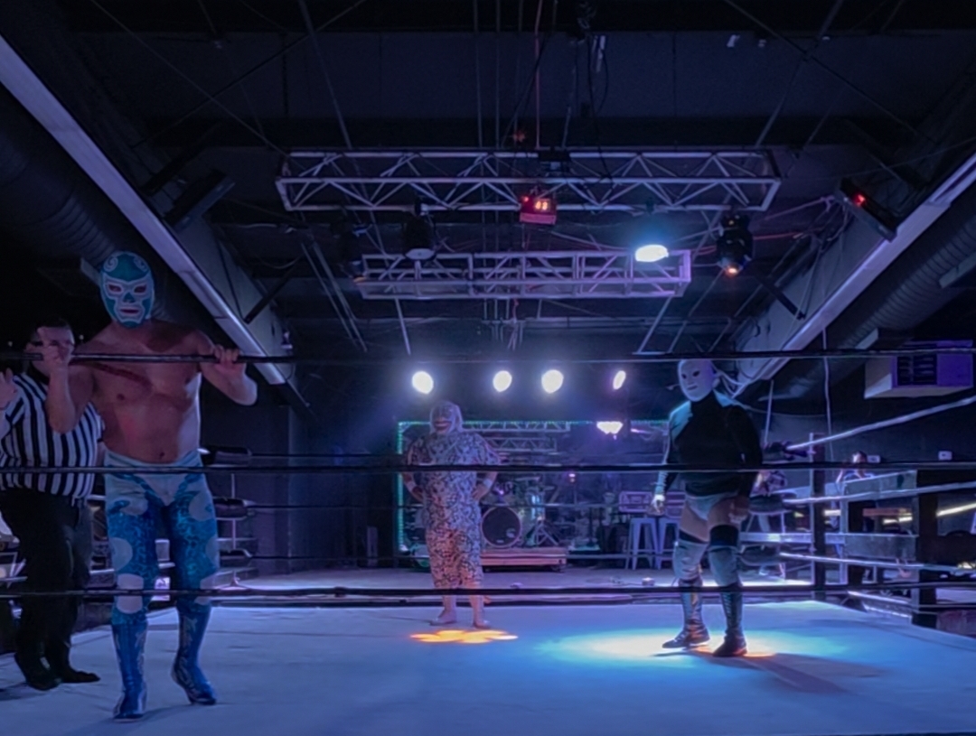 Wrestlers and referee in a wrestling ring with bright overhead lights and audience area.