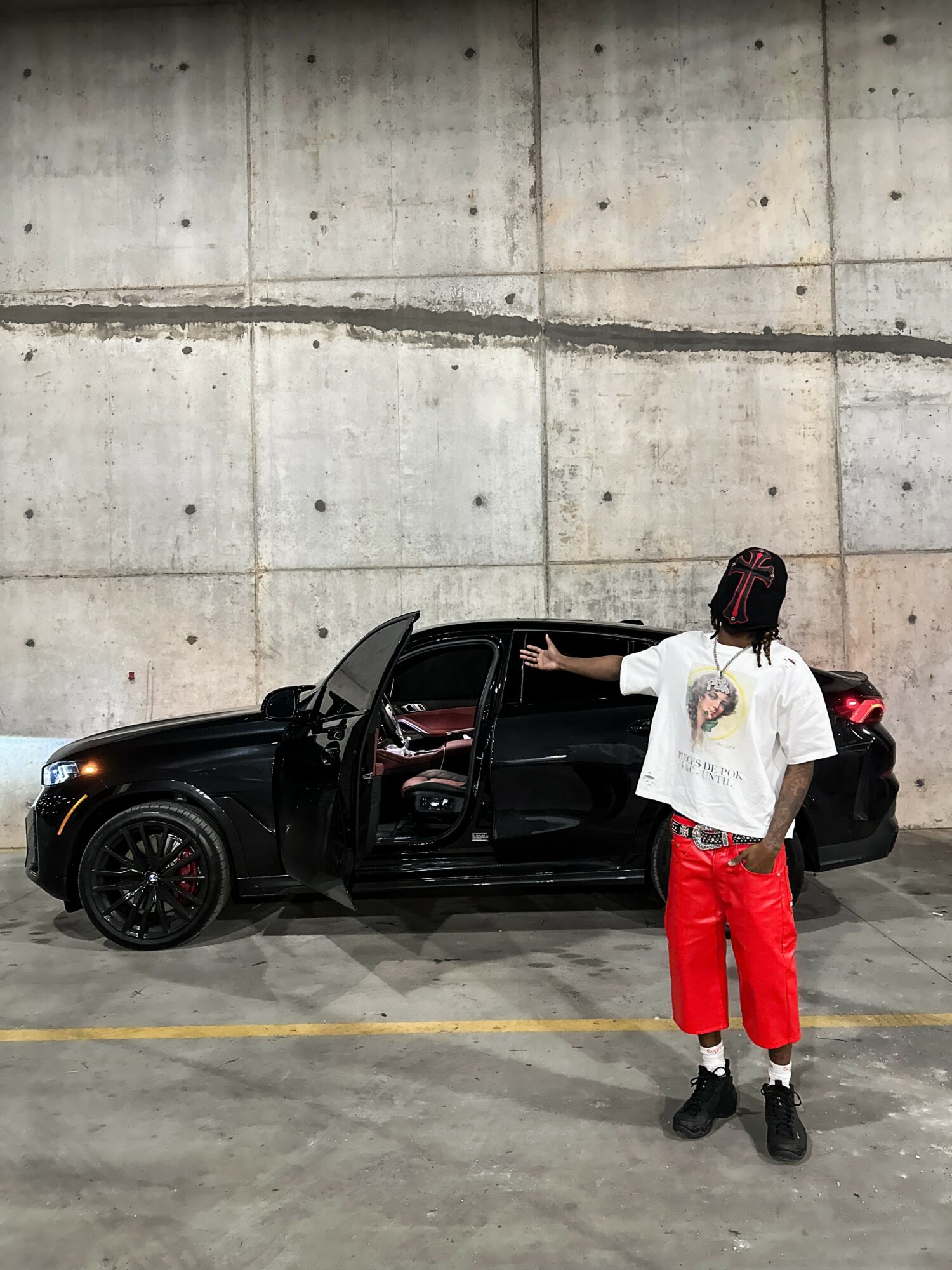 Person with braided hair, white t-shirt, red pants, black shoes, pointing at black car in parking garage.