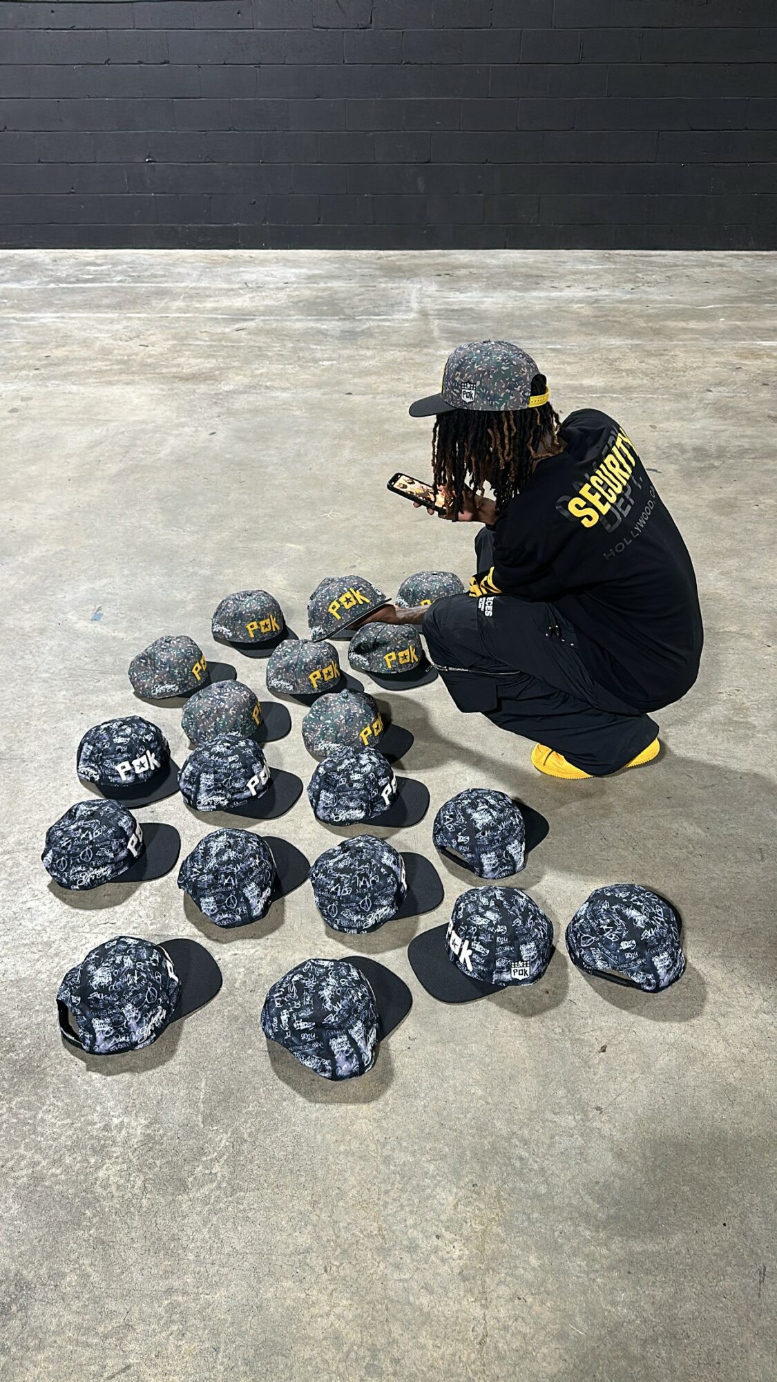 Person crouching on ground, wearing a black jacket with yellow text, gray hat, surrounded by rocks with white markings.