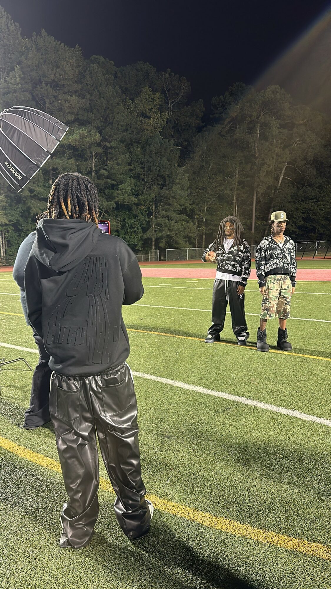 Person with dreadlocks wearing a black jacket and shiny pants stands on a field at night, facing two people in camouflage and black jackets.