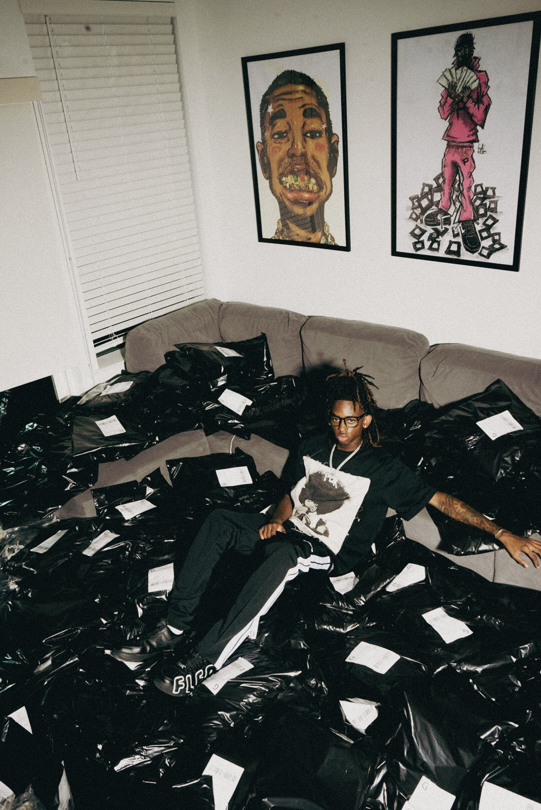 Person sitting on a couch surrounded by numerous black bags, holding a book, in a room with framed artwork on the wall.
