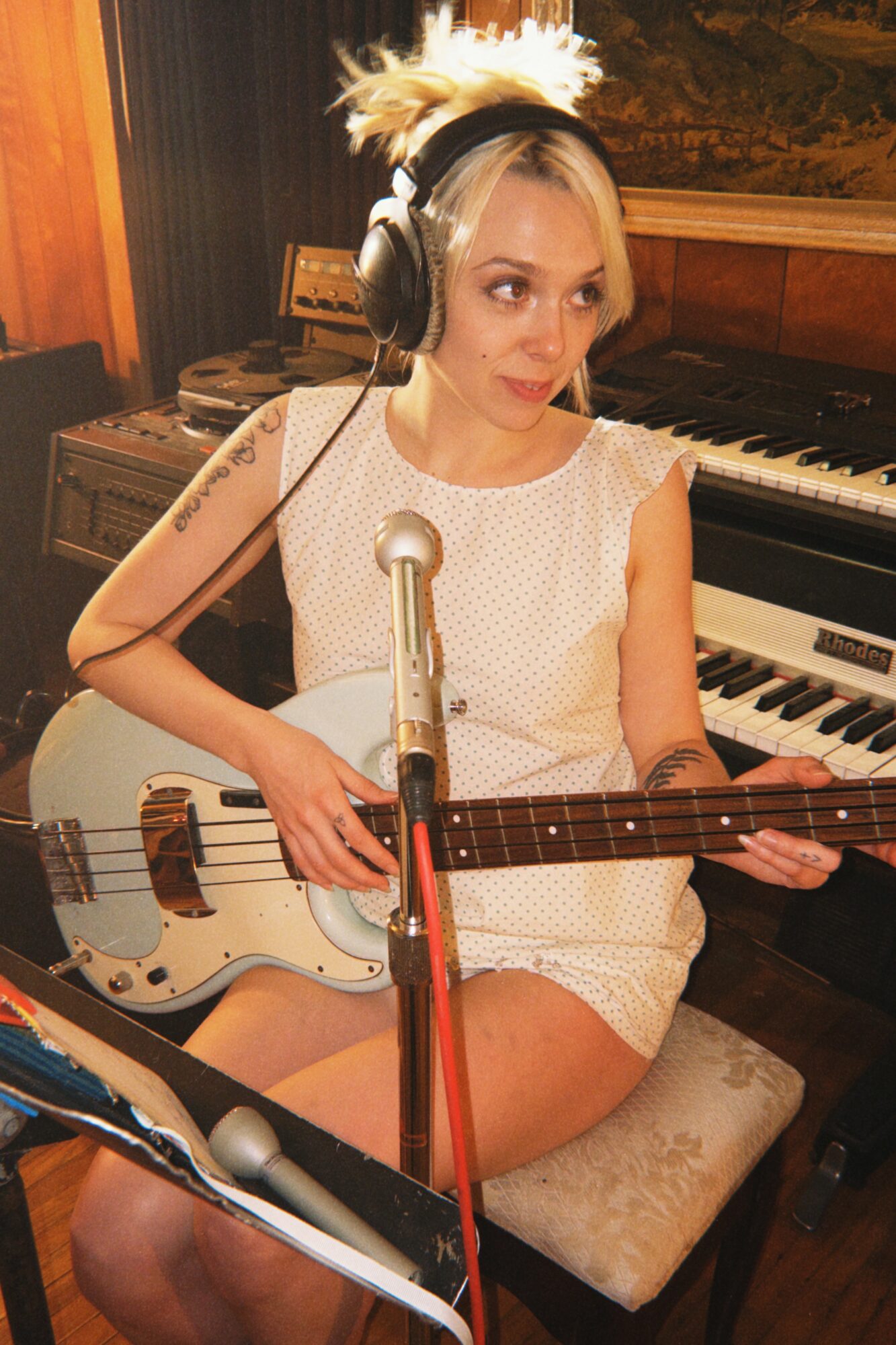 Young woman with blonde hair wearing headphones, sitting on a chair, holding a guitar, in a music studio.