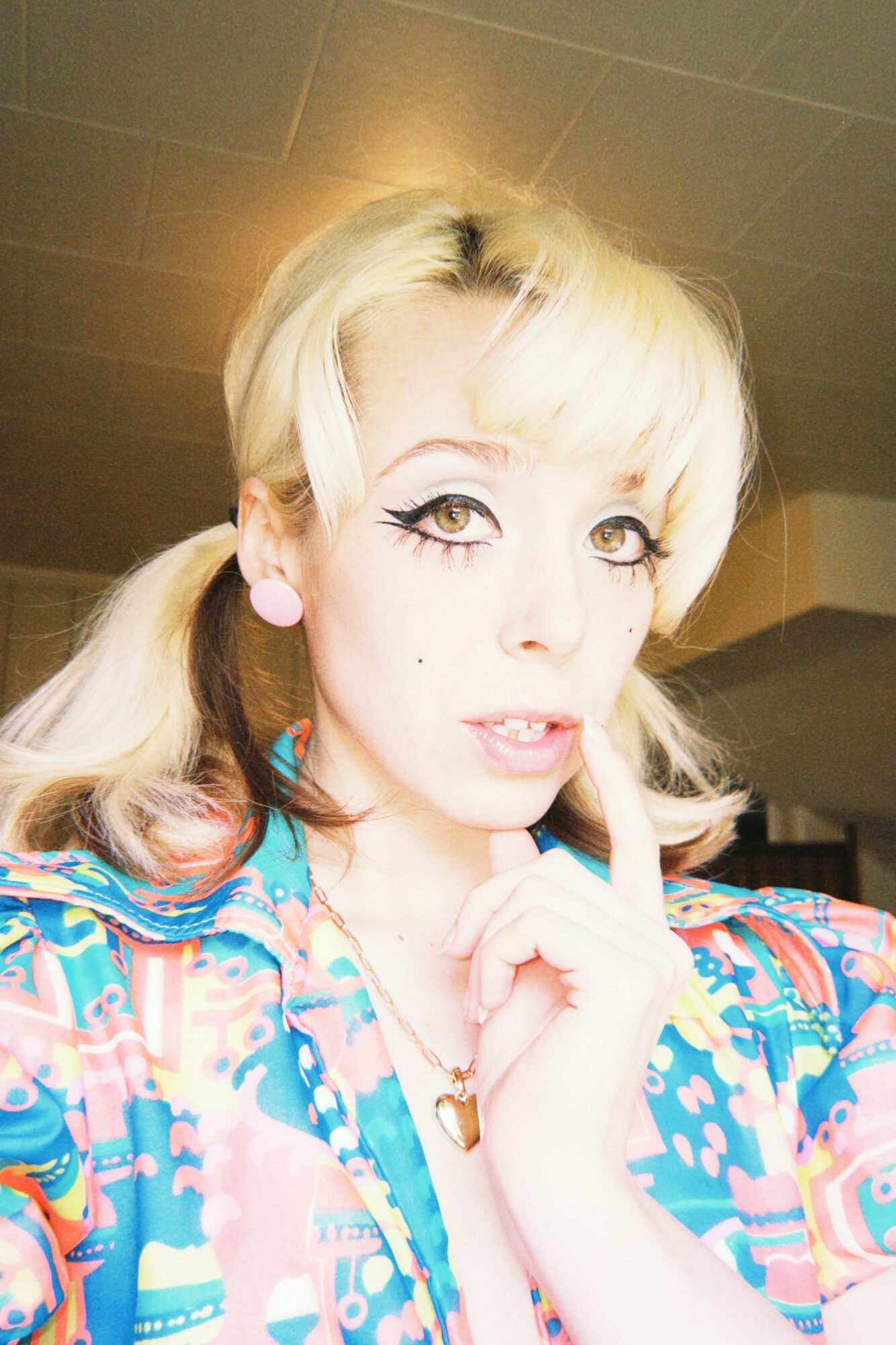 Woman with blonde hair, makeup, and colorful patterned clothing, holding her finger to her lips.