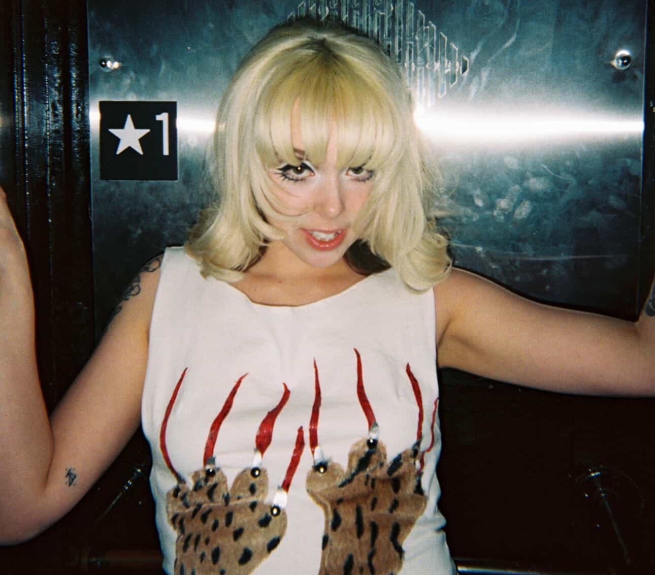Woman with blonde hair wearing a white top with animal paw prints, posing with arms outstretched.
