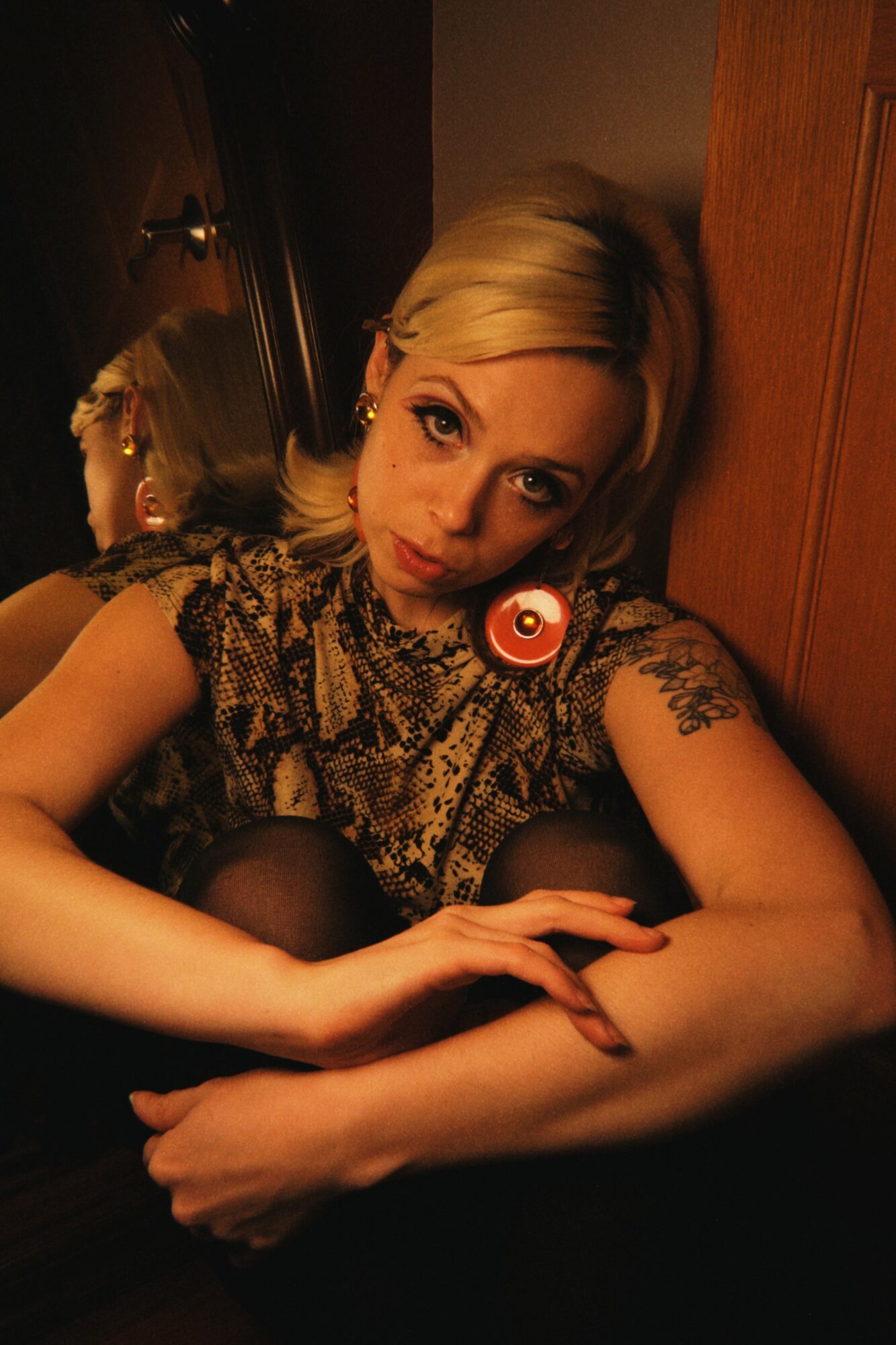 Woman with blonde hair and tattoos resting her arms on a surface, looking at the camera in a dimly lit room.
