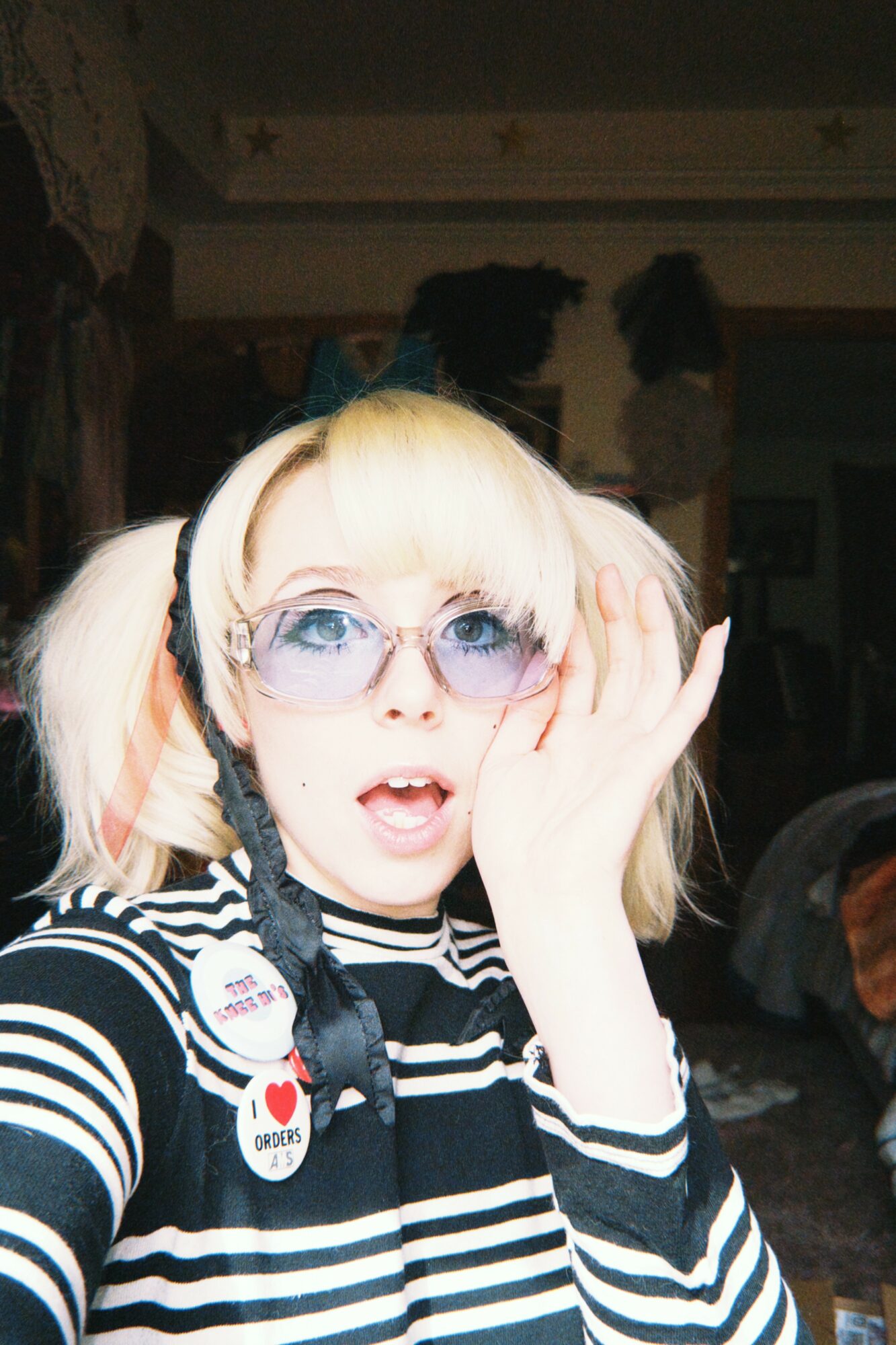 Person with blonde hair, wearing sunglasses, striped shirt, and accessories, holding hand to face with surprised expression.
