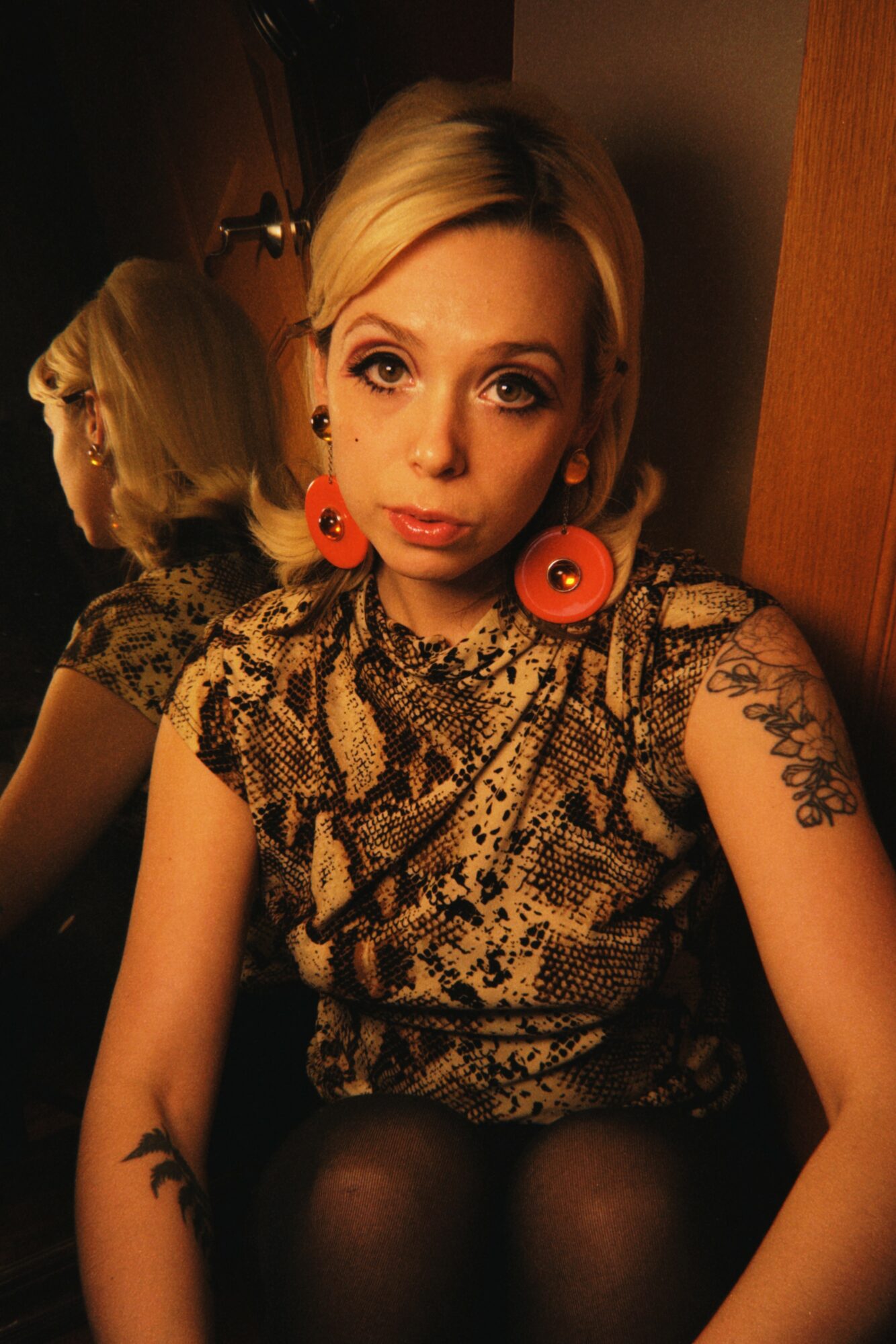 Young woman with short blonde hair wearing large orange earrings and a snake print top, sitting indoors.
