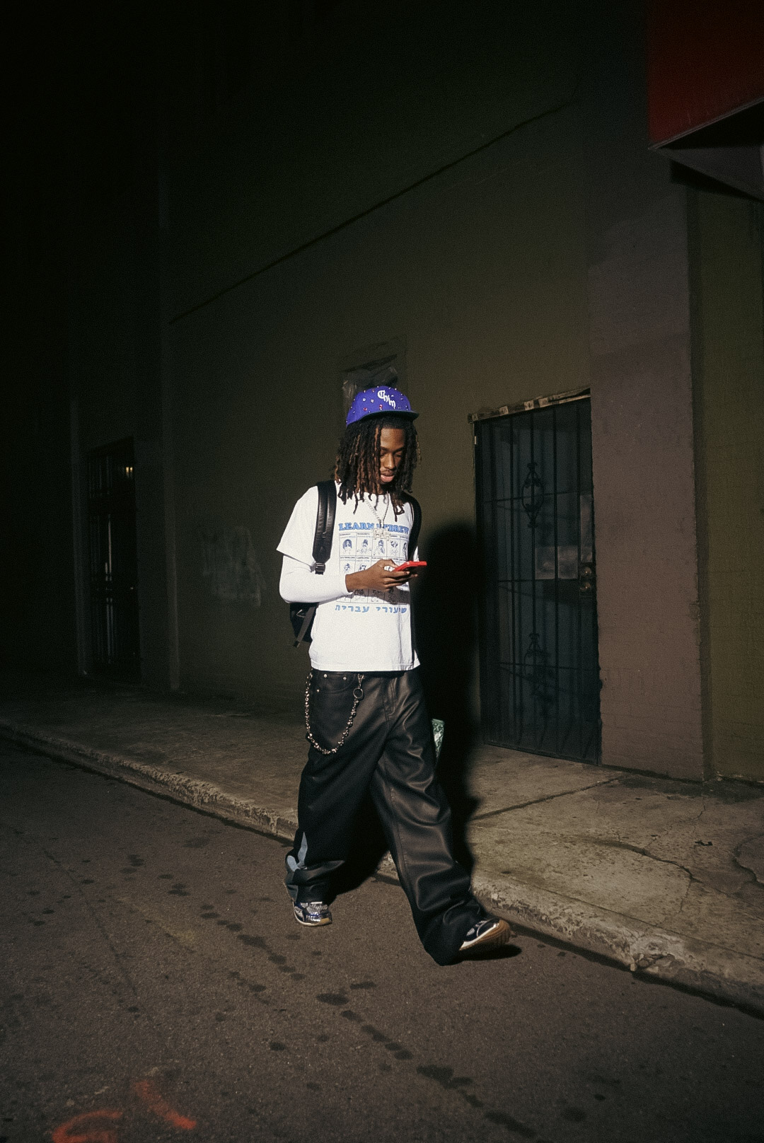 Person walking on sidewalk at night, wearing a hat, white shirt, and dark pants, holding a phone.