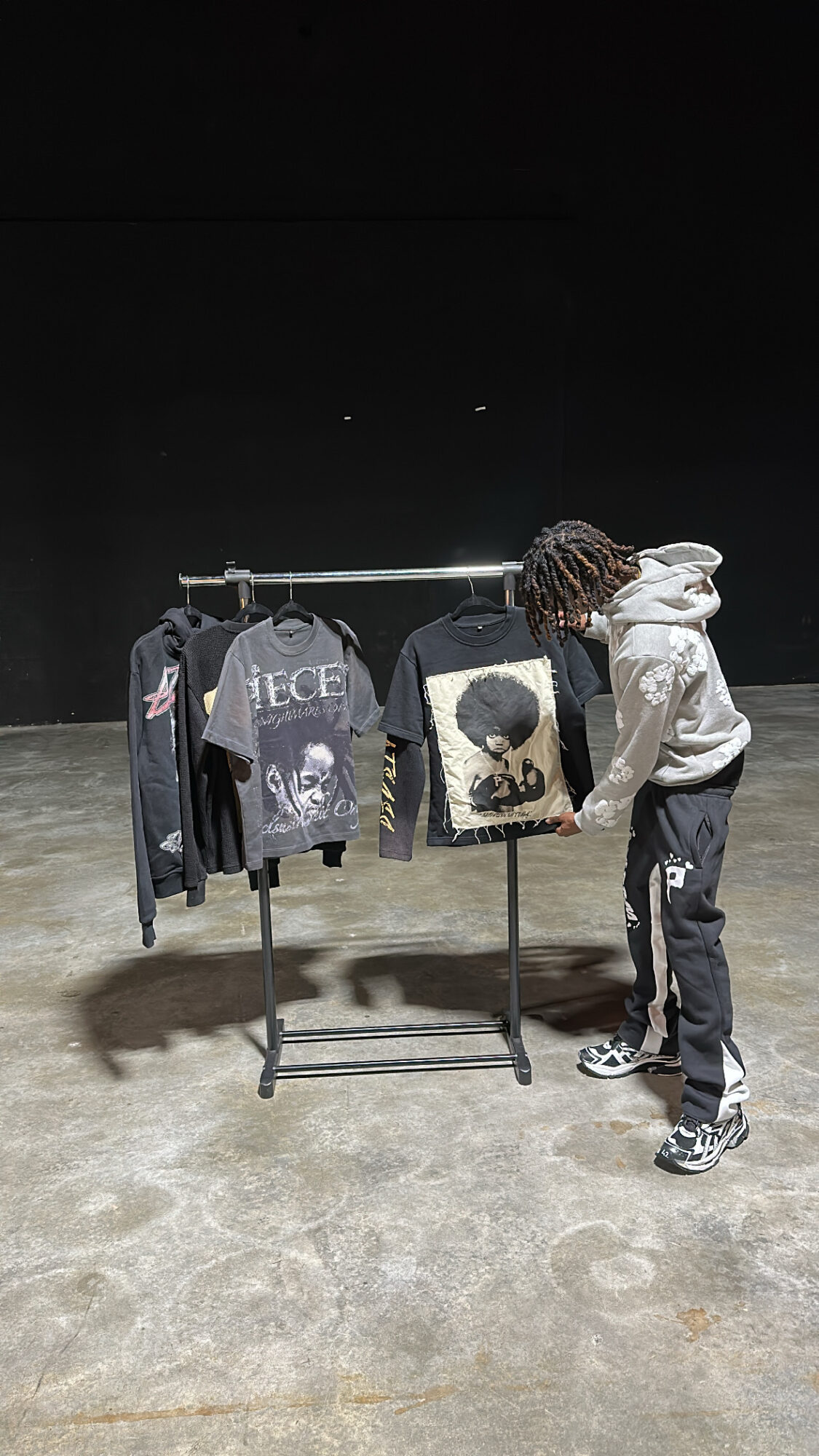 Person with dreadlocks wearing a white hoodie and black pants looking at clothing displayed on a rack in a dark space.