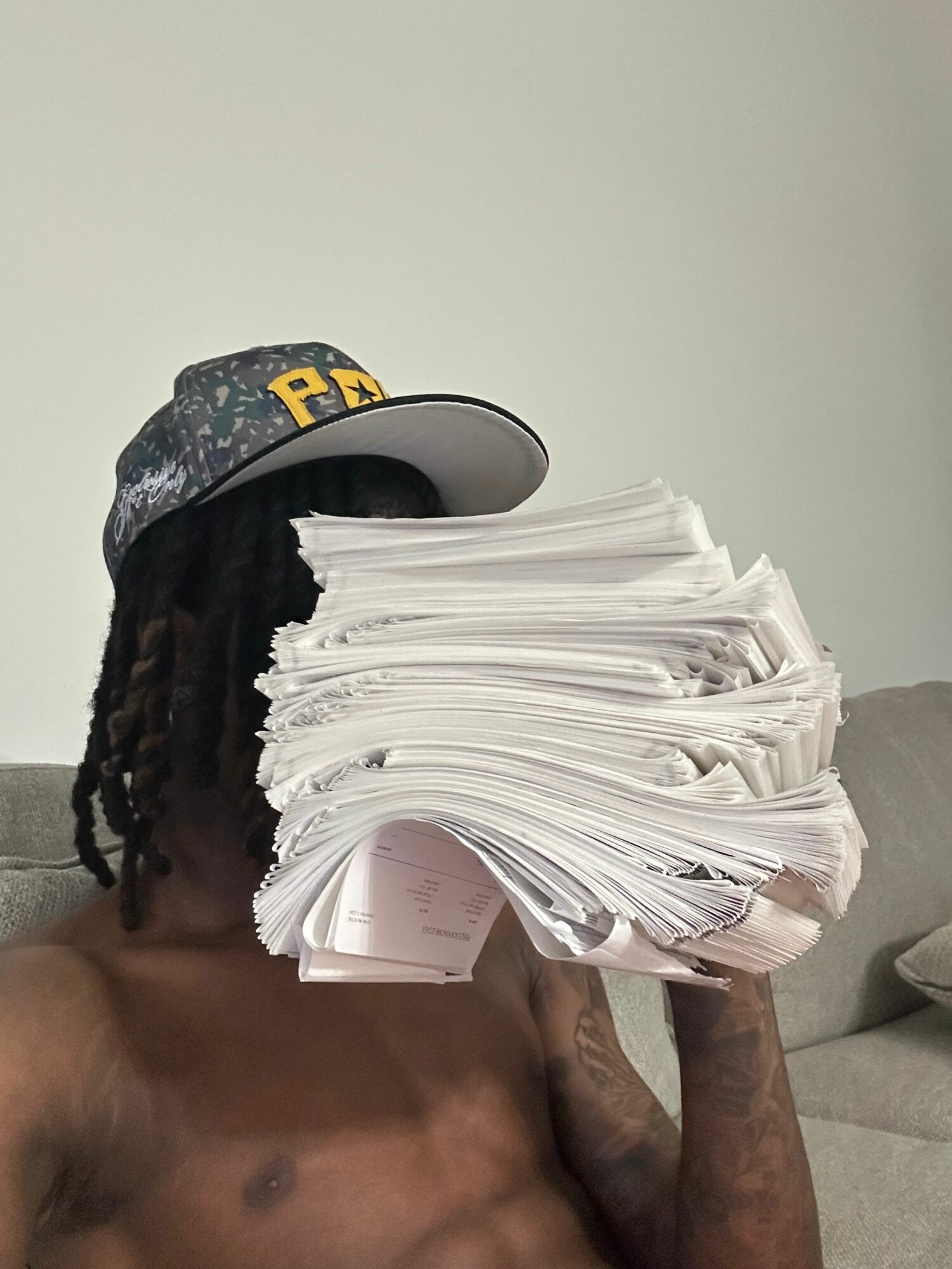 Person wearing a cap with yellow letters, holding a large stack of papers in front of their face, shirtless.