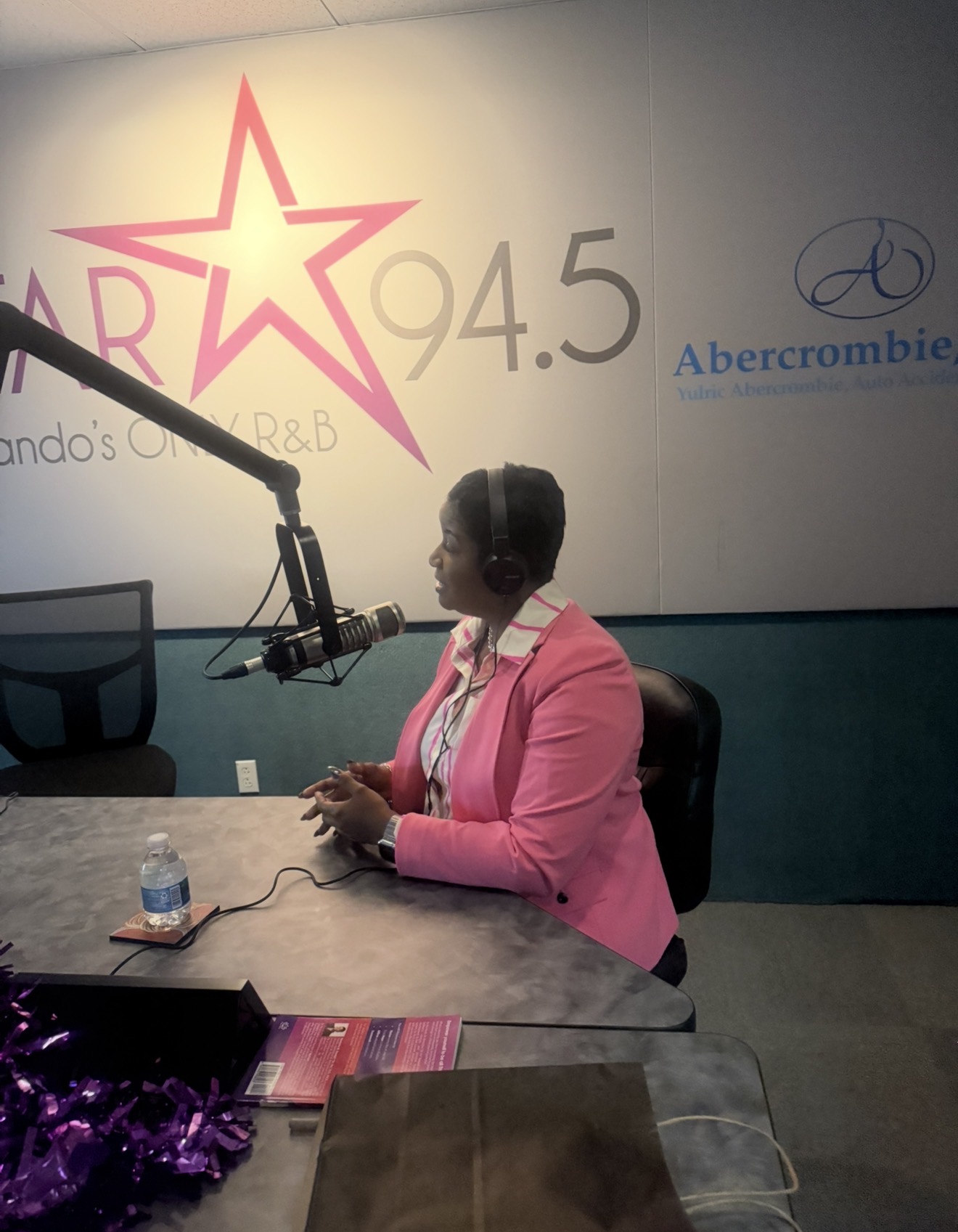 Woman wearing pink blazer and headphones speaking into microphone at radio station studio.