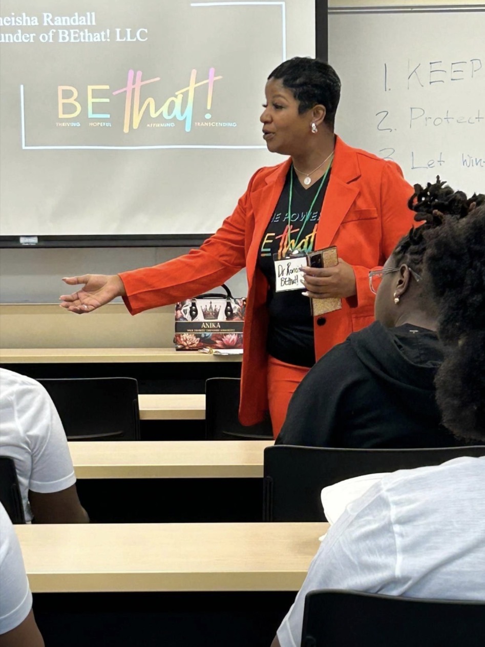 Woman in red blazer speaking to classroom, gesturing with hand, students listening attentively.