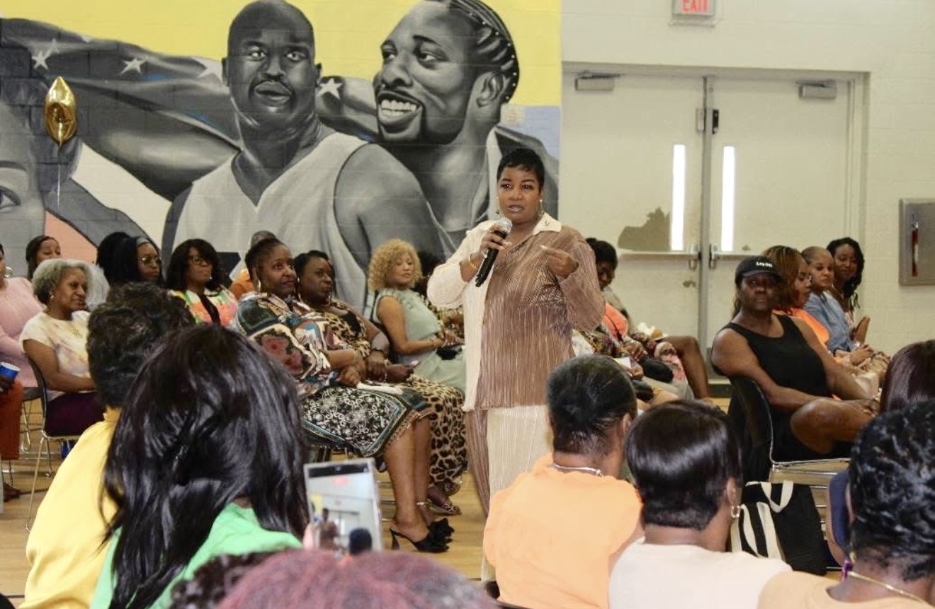 Person speaking into microphone in front of seated audience in a room with mural of two men on wall.