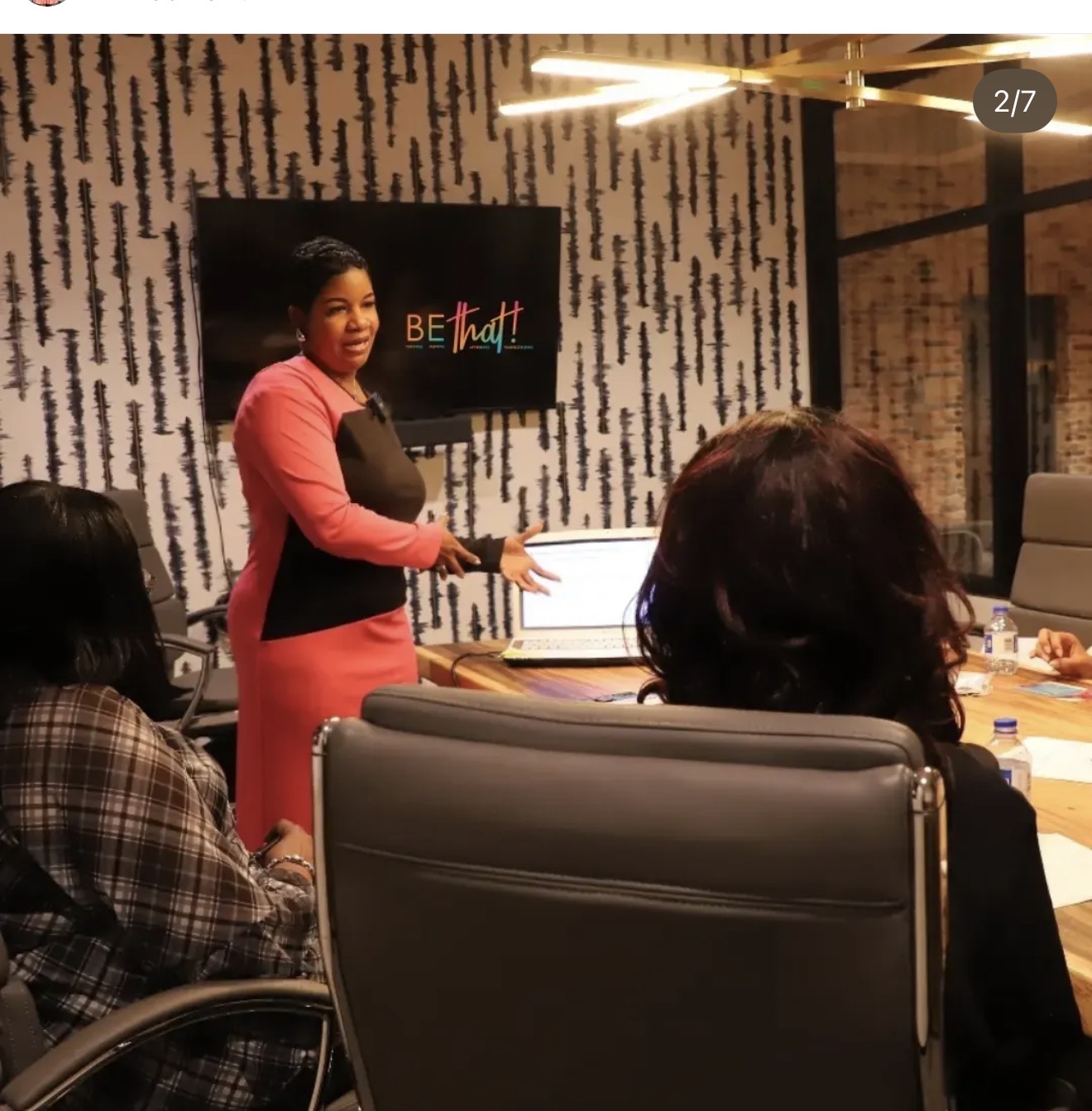 Woman in pink blazer standing and presenting to seated group in a conference room.