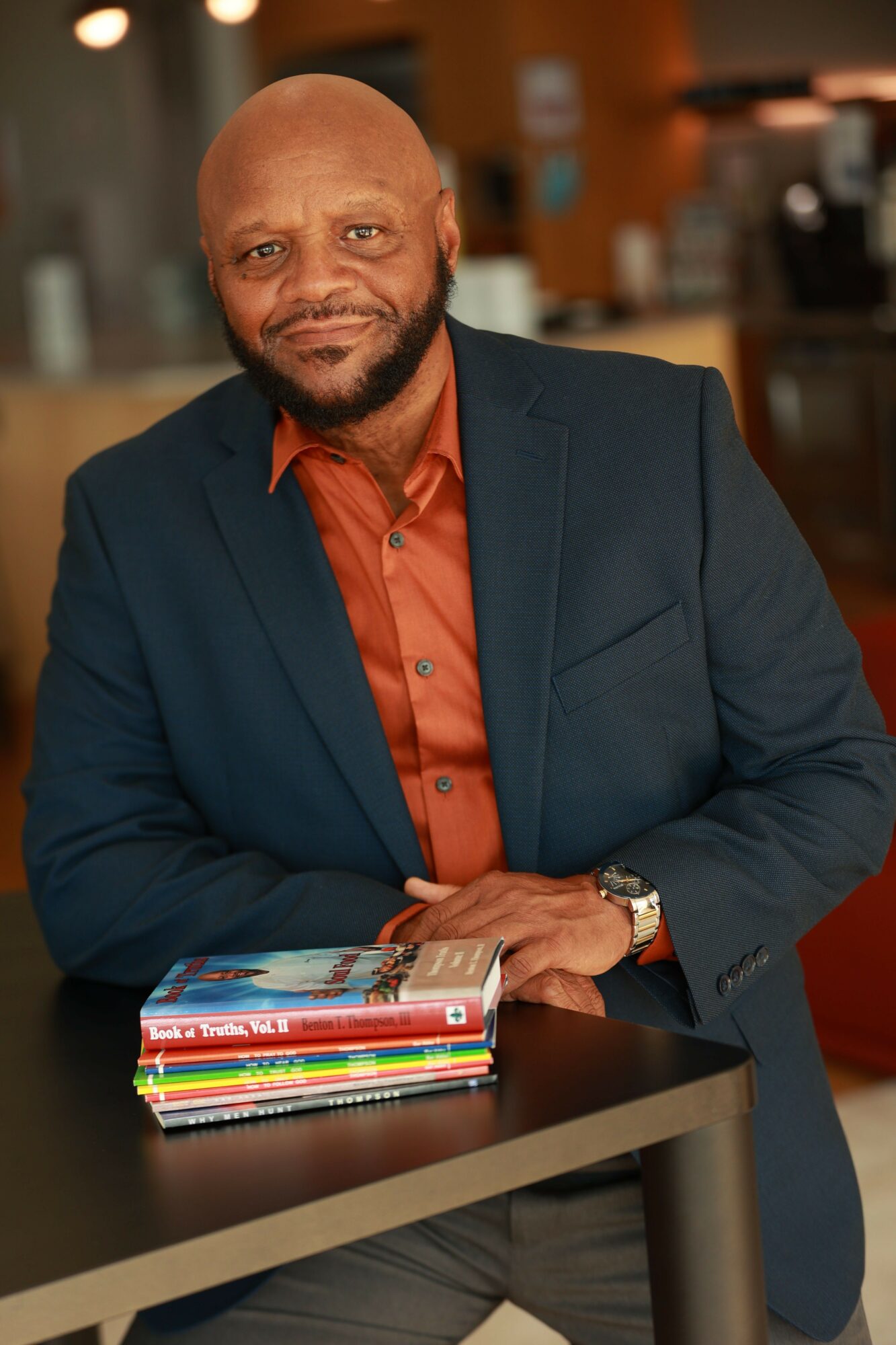 Man with a beard and bald head wearing a dark blazer and orange shirt, standing at a table with magazines.