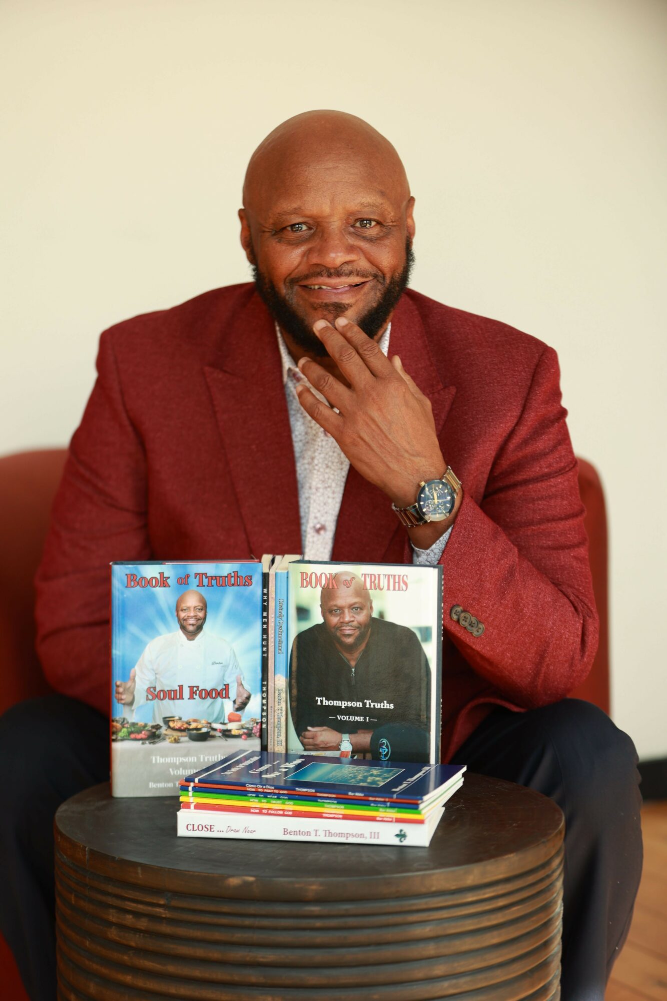 Man with a beard and bald head, wearing a red blazer, sitting at a table with books and smiling.