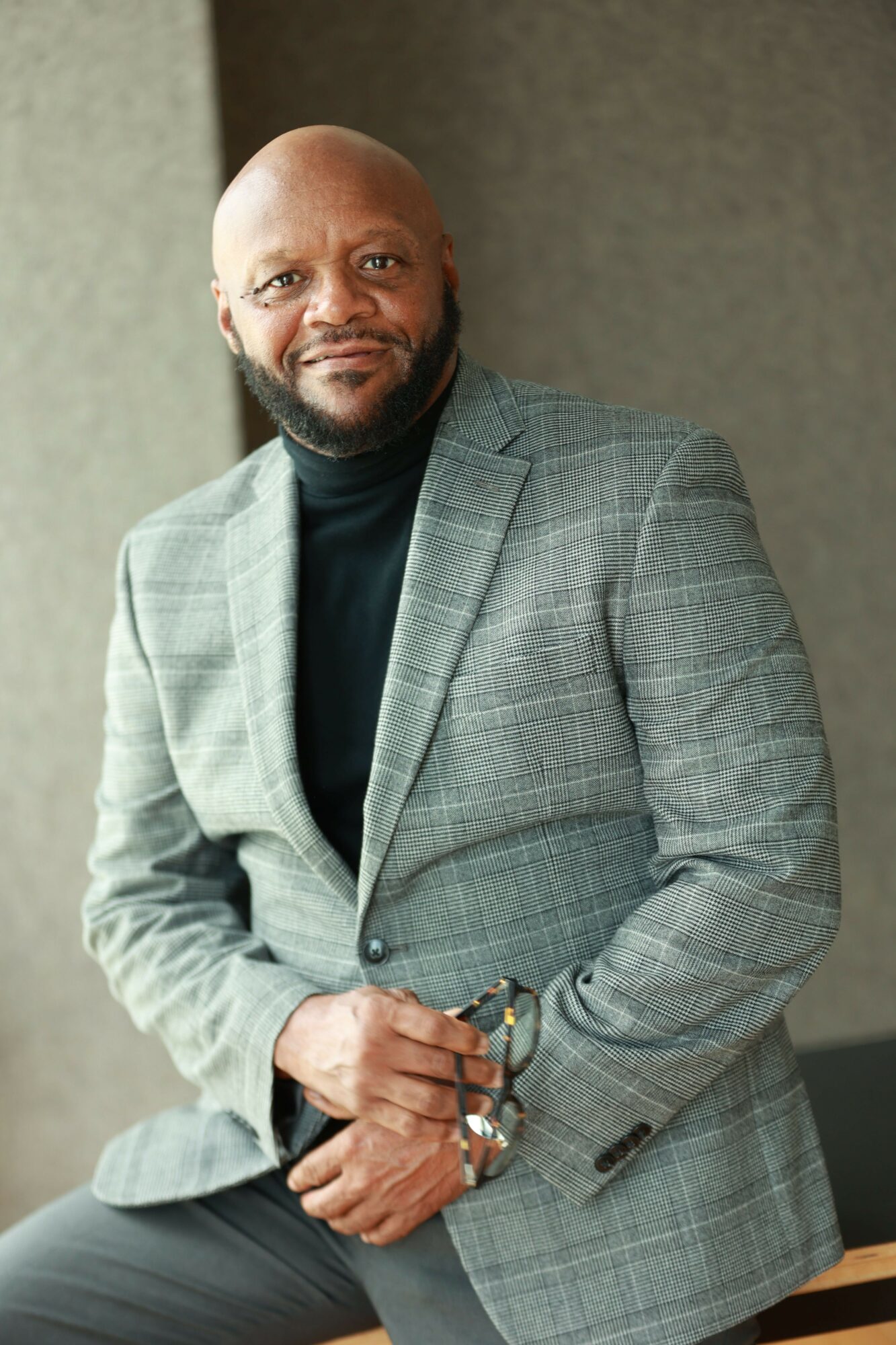 Man with a beard in a gray checkered blazer and black turtleneck, holding glasses, standing indoors.