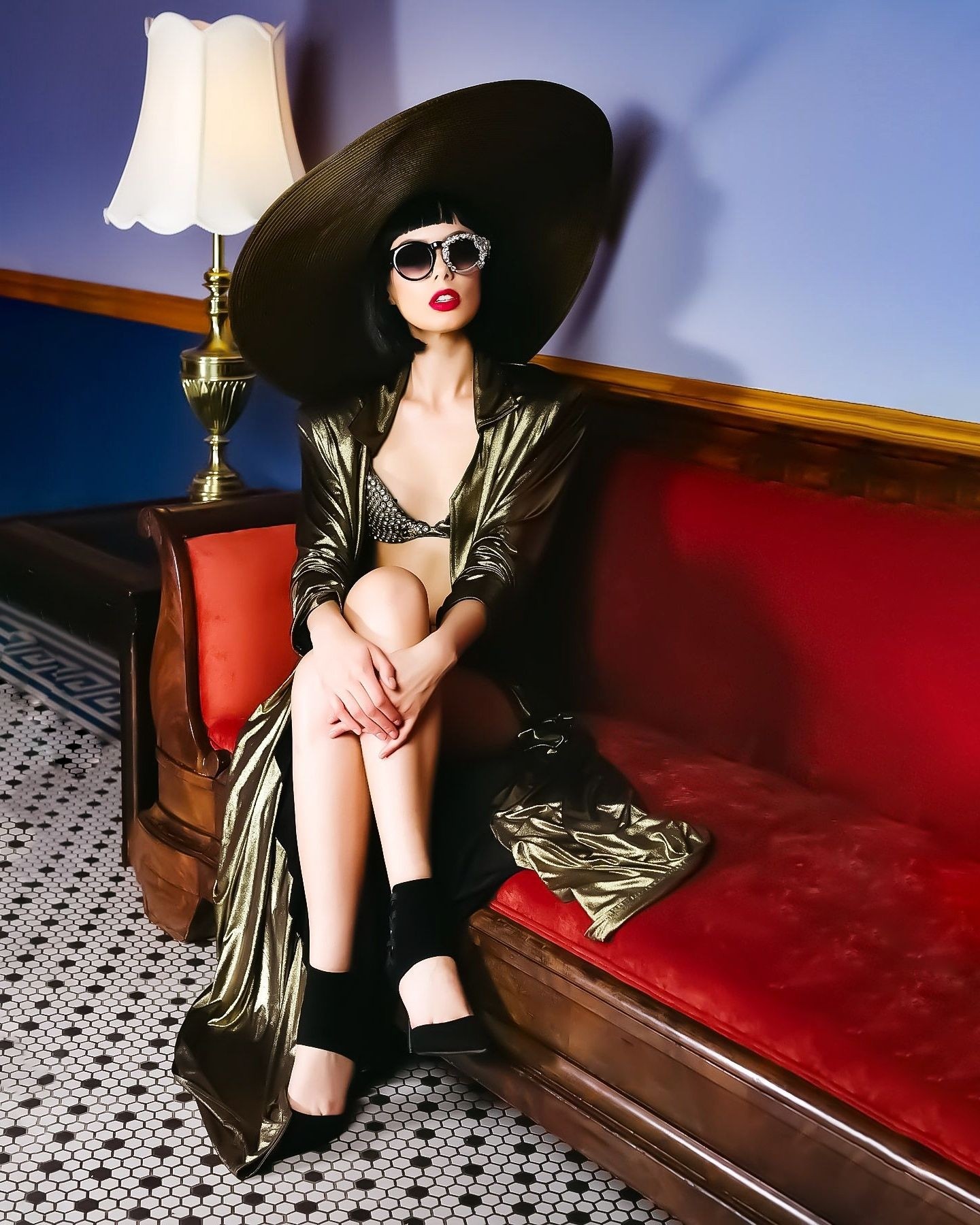 Woman sitting on a red velvet sofa, wearing large hat, sunglasses, and shiny black outfit, in a room with a lamp and patterned floor.