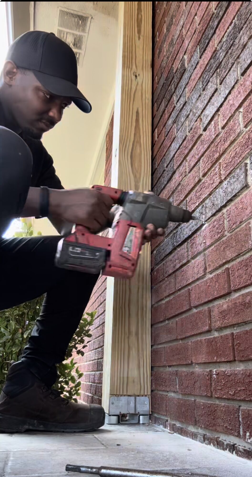 Person using a power drill on a brick wall outdoors, wearing a black cap and dark clothing.