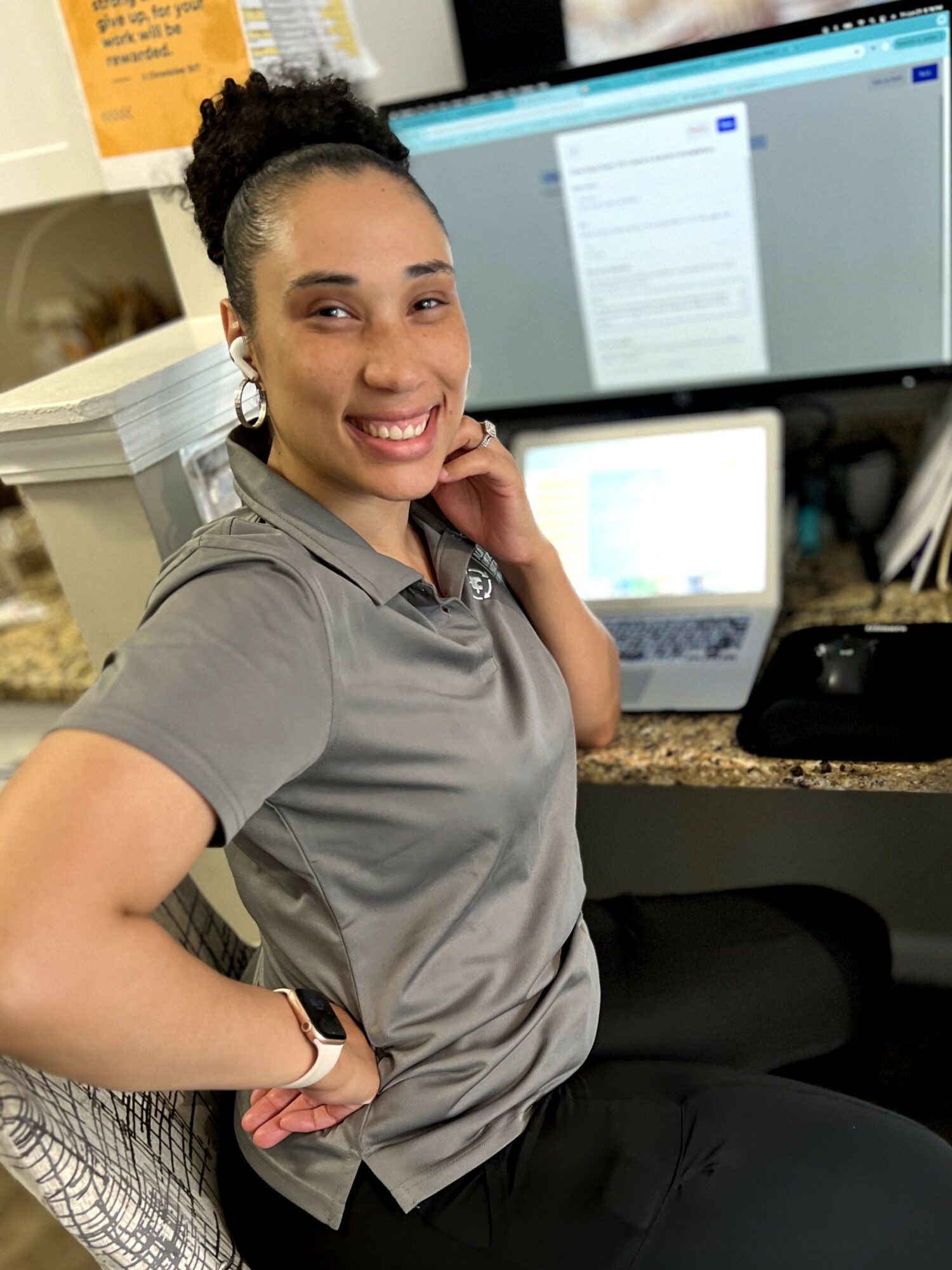 Woman smiling, sitting at desk with computer monitors, wearing gray polo shirt, in office environment.