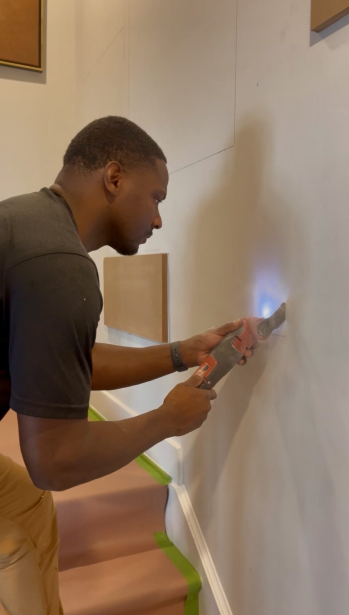 Person using a tool on a wall in a room with framed pictures, beige wall, and wooden furniture.