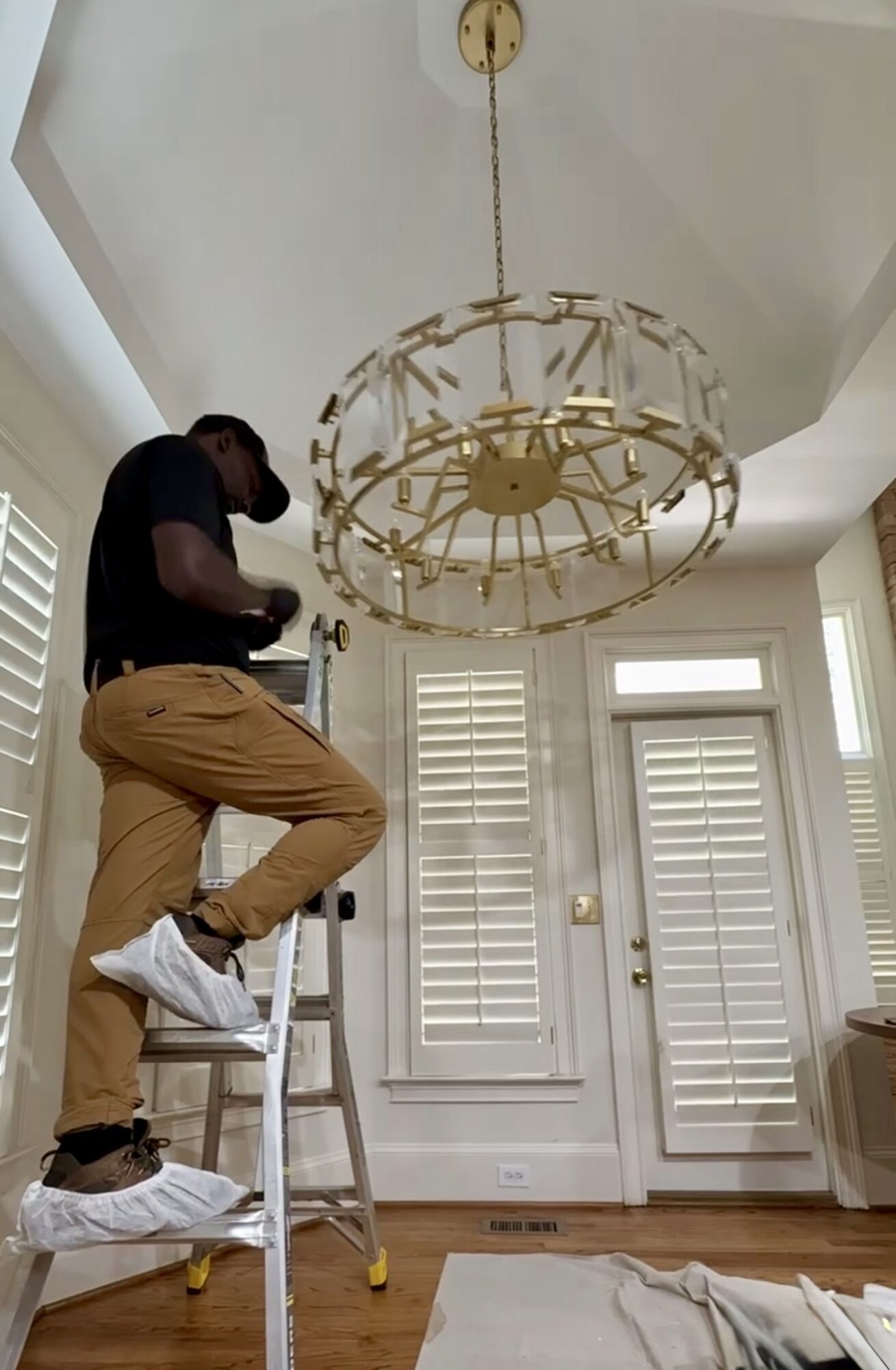 Person standing on a ladder adjusting a large, gold, circular chandelier in a room with white shutters and a door.
