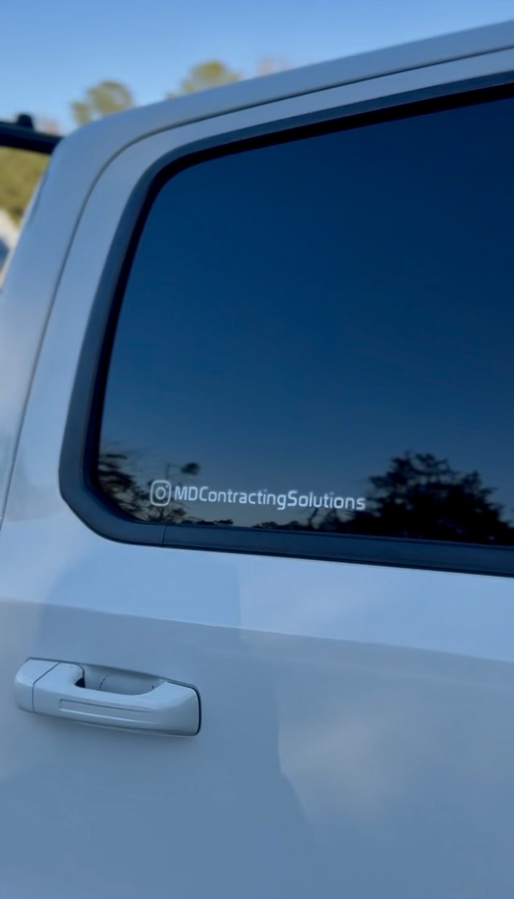 Close-up of a car window with a sticker reading '@MDContractingSolutions' and a reflection of trees and sky.