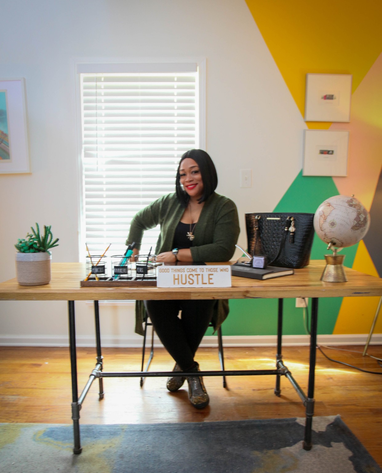 Woman sitting at desk with a sign that says 'HUSTLE', surrounded by plants, a globe, and office supplies.